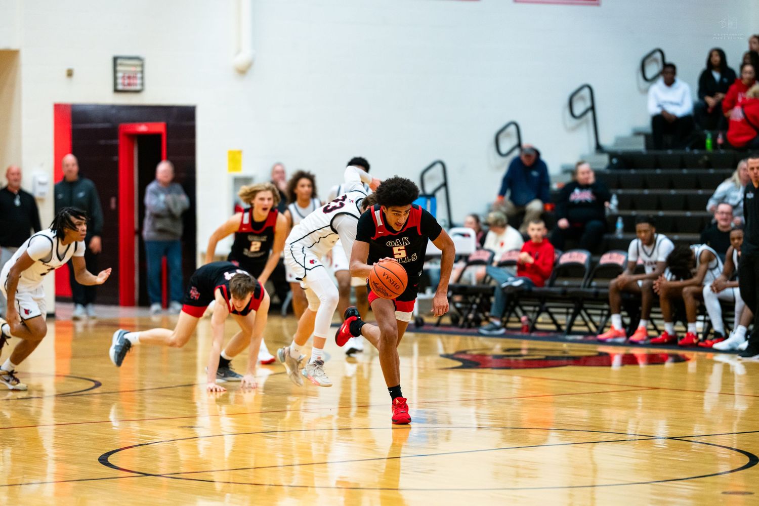 Players scramble for loose ball during intense high school basketball game in red and white gym.