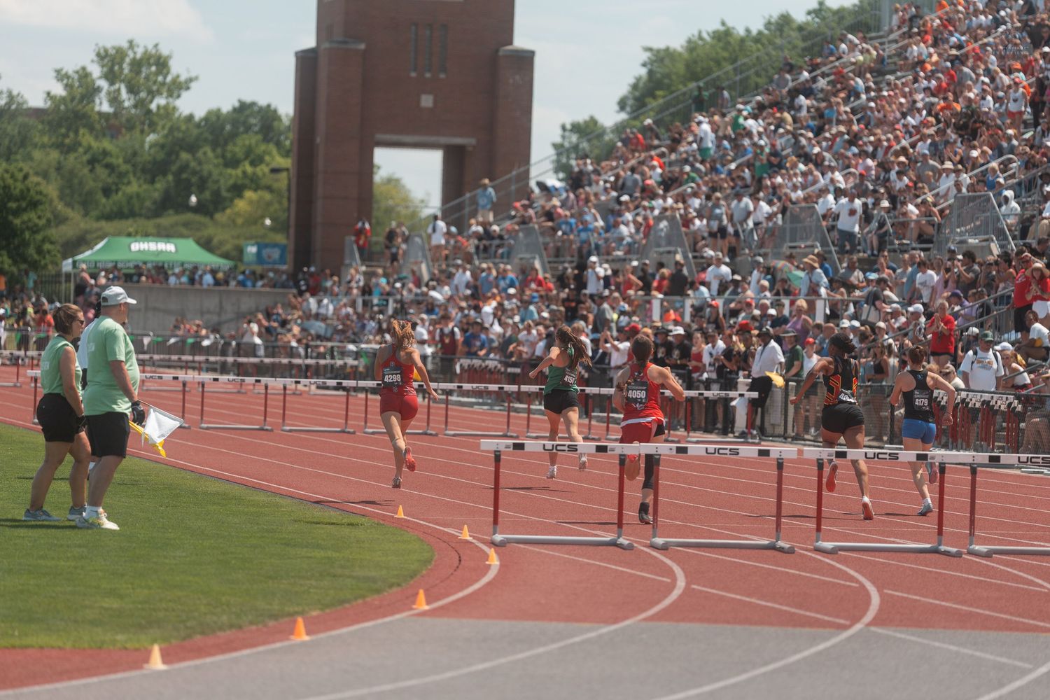 High school athletes clear hurdles on a red track with a large crowd watching from the grandstands behind them.