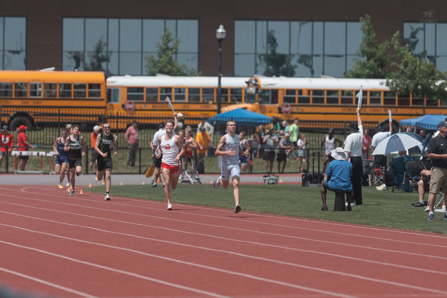 Track athletes race on a red track with yellow school buses visible in the background on a sunny day.