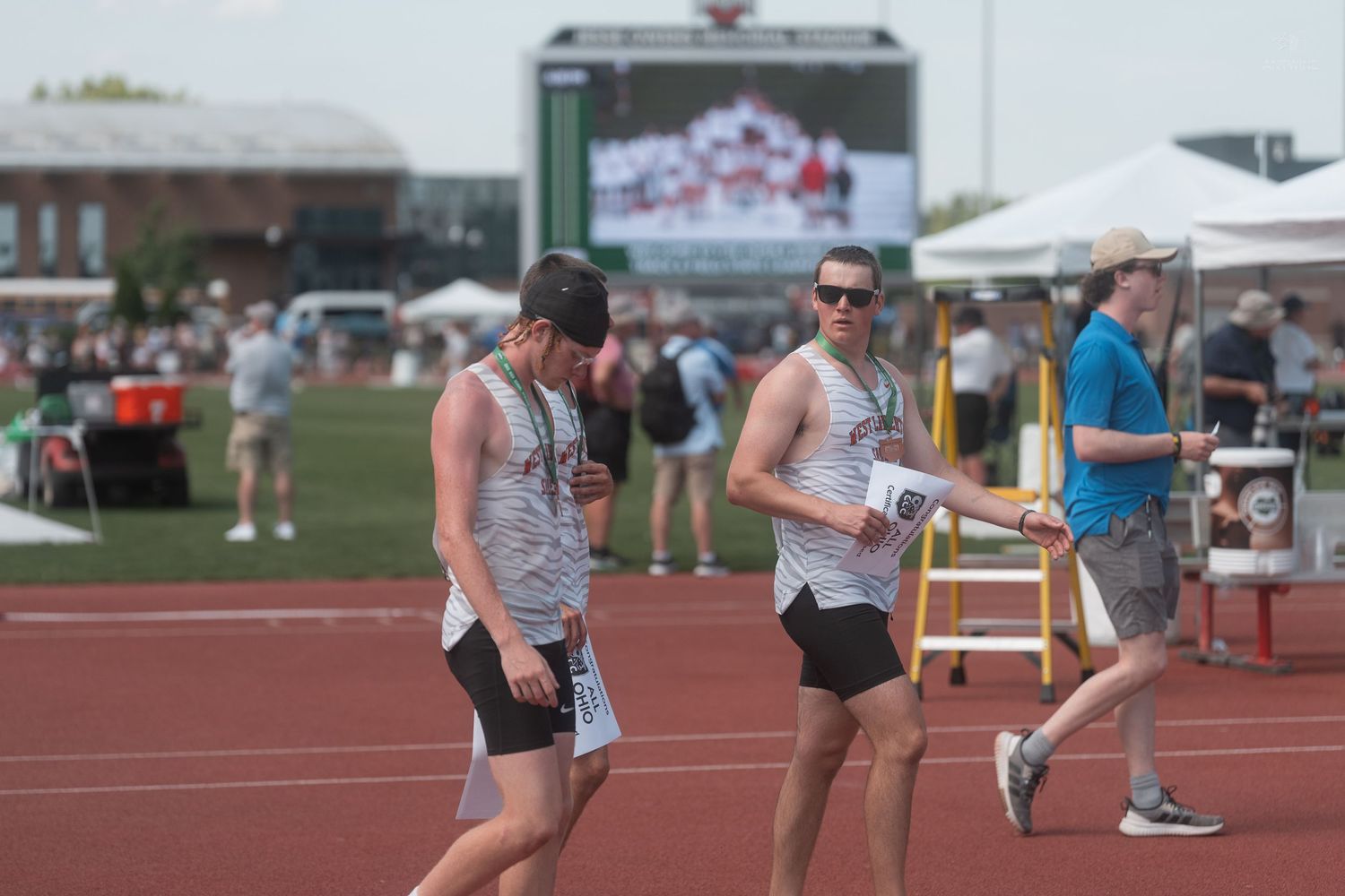 Athletes walk on a track field during an outdoor track and field competition with a large video screen in the background.