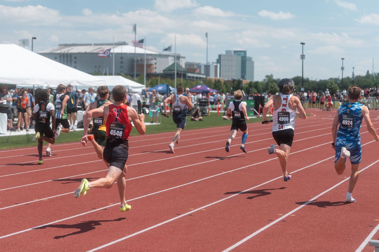 Runners compete in a sprint race on a red outdoor track during a sunny athletic meet with spectators lining the field.