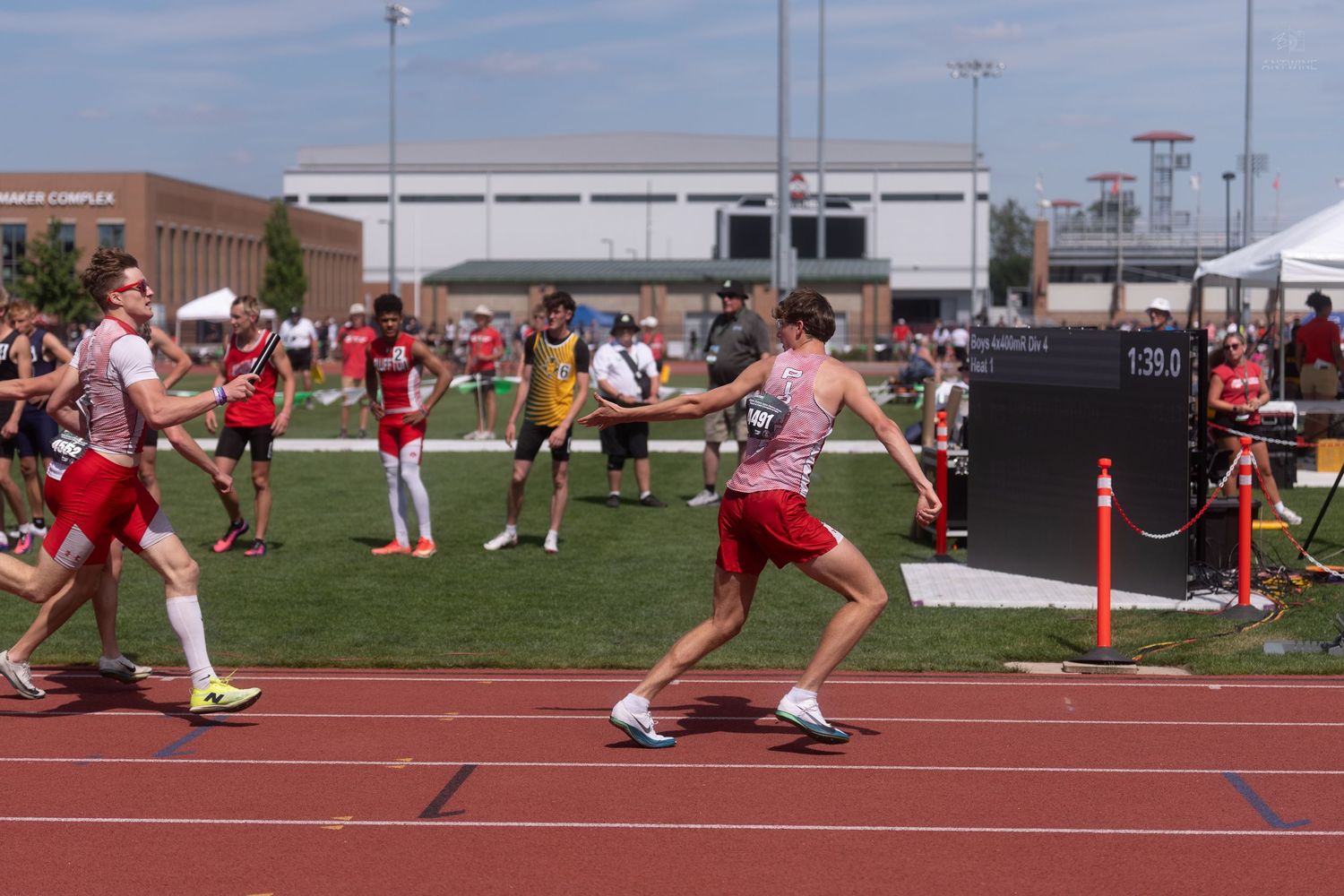 A relay runner reaches out to receive a baton handoff during an outdoor high school track championship.