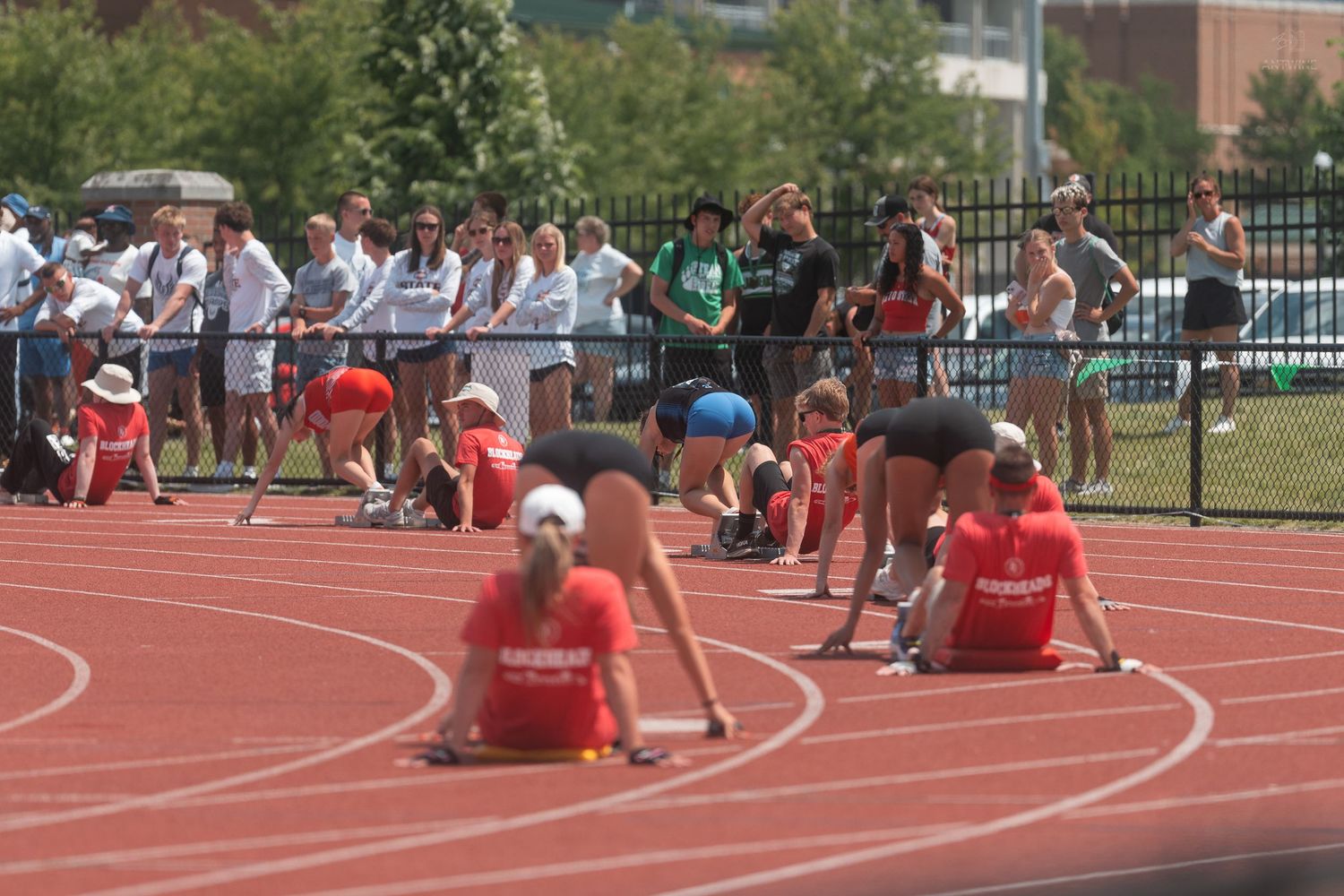Athletes crouching in starting positions on red track at outdoor collegiate track and field meet with large crowd.
