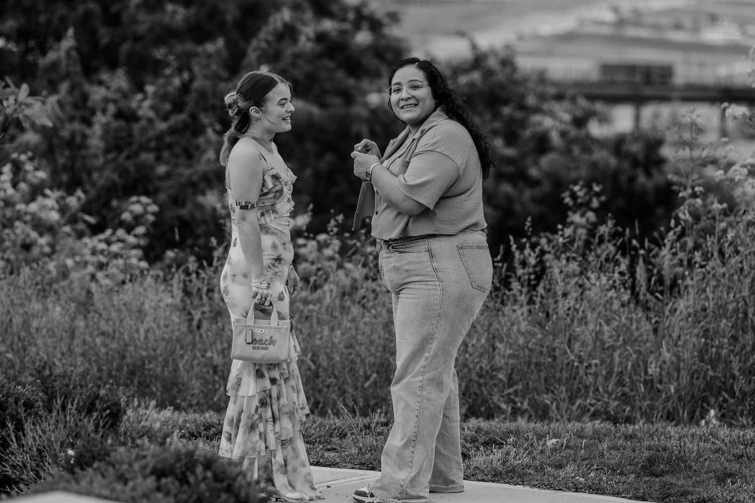 Two women share a joyful conversation outdoors surrounded by lush greenery in black and white photo.