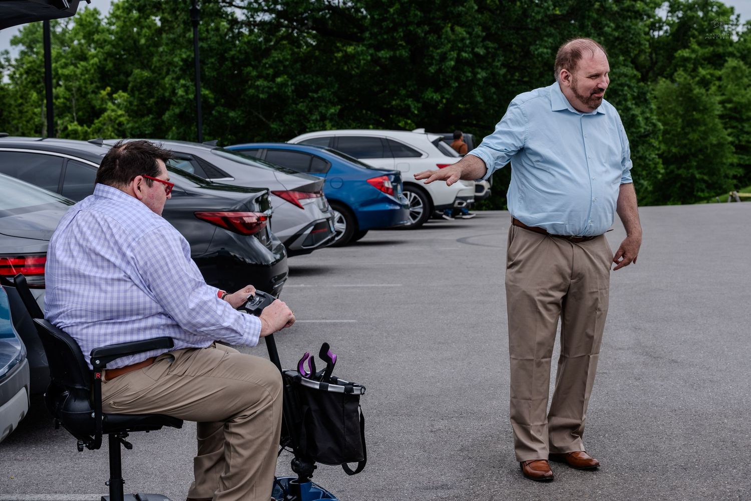 Man in blue shirt pointing at another man seated on a mobility scooter in a parking lot.