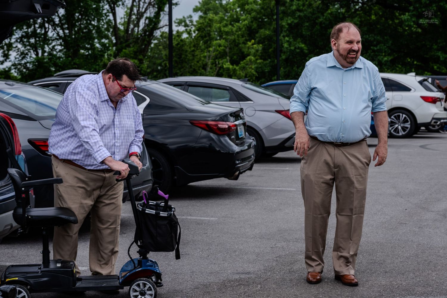 Two men in a parking lot, one seated on a mobility scooter and one standing in a blue shirt.