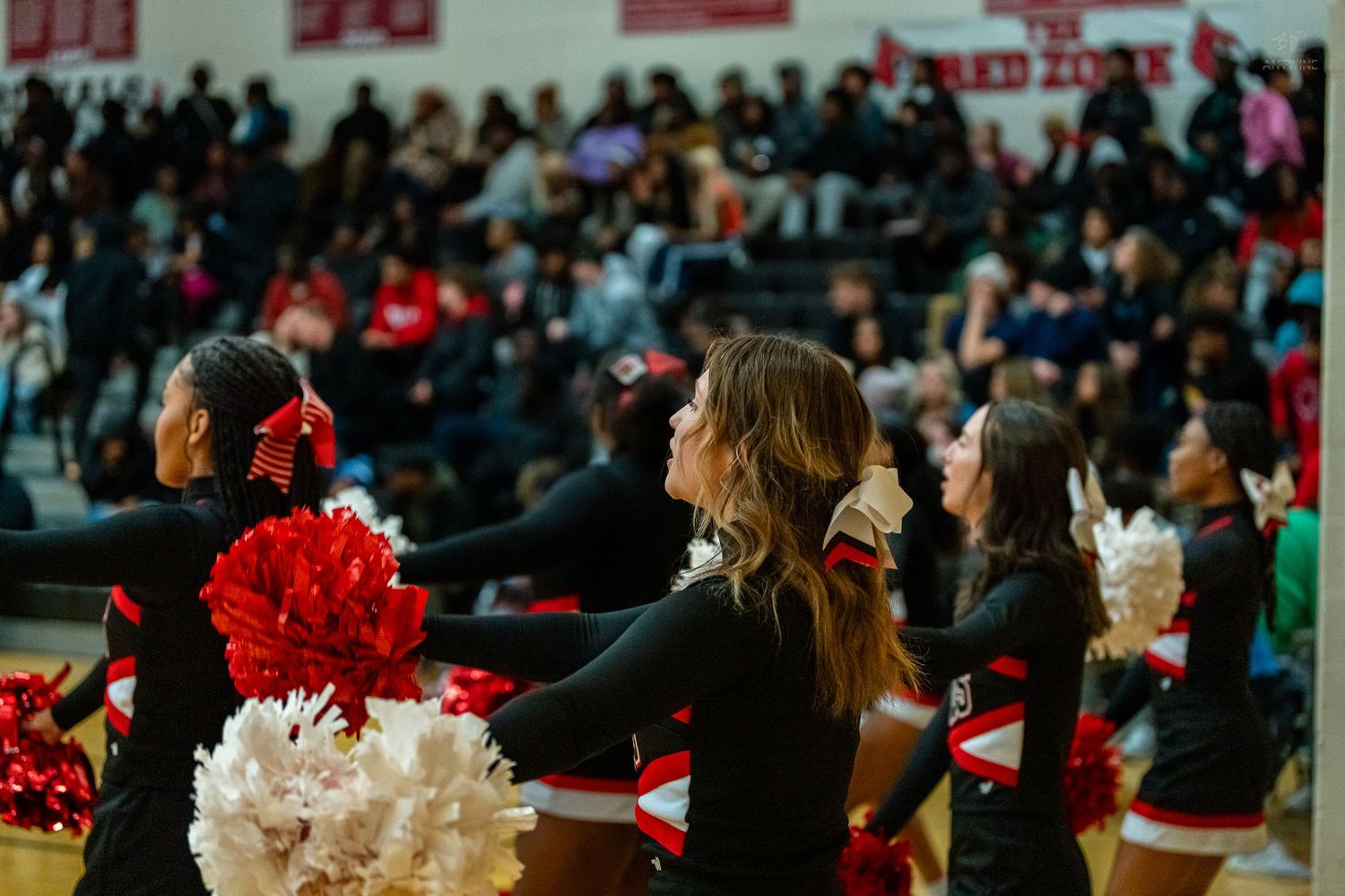 High school cheerleaders in black uniforms wave red and white pom poms before packed gymnasium crowd.