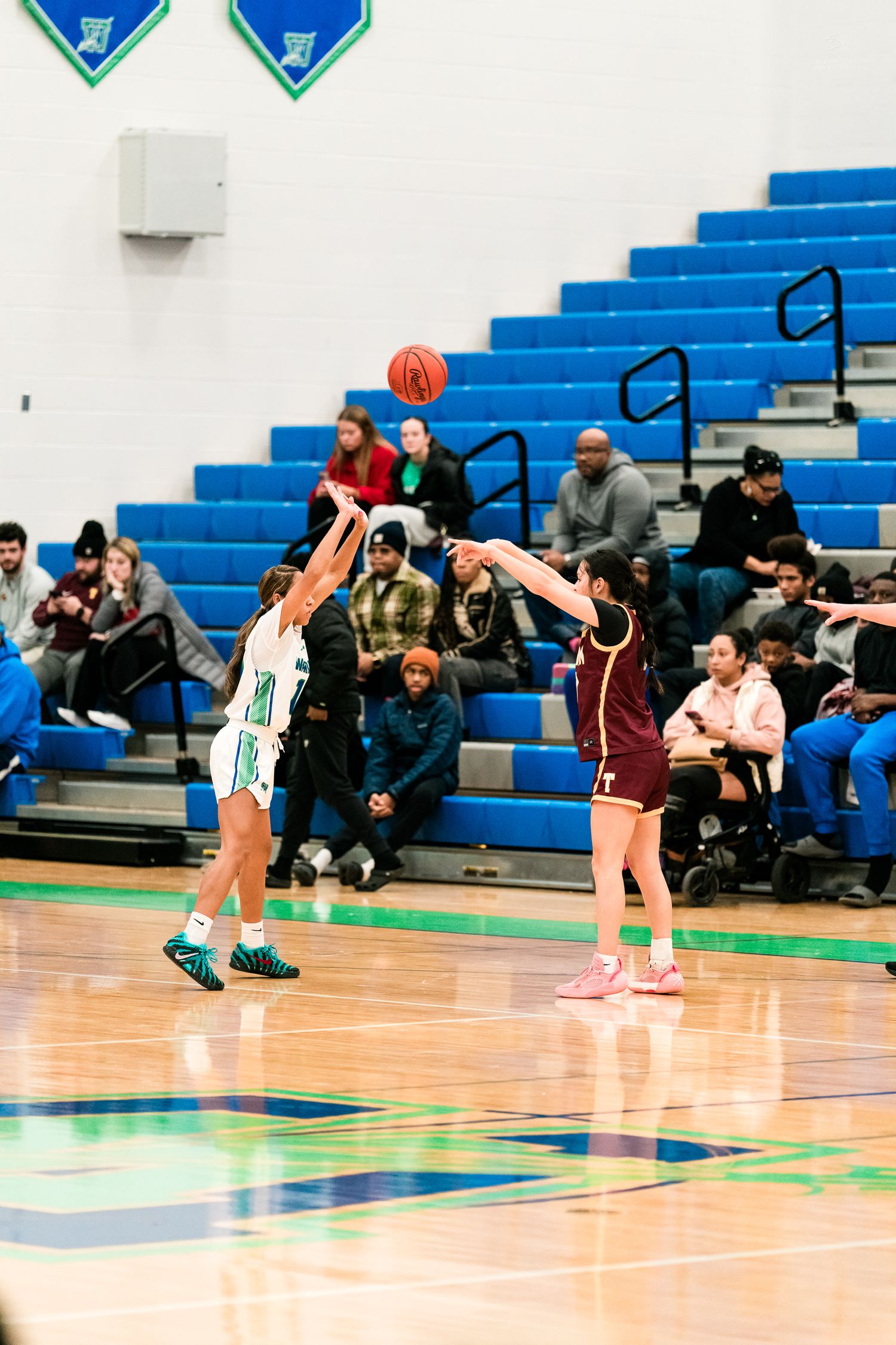 Female basketball player in teal uniform shoots over a maroon-uniformed defender during a high school girls basketball game.