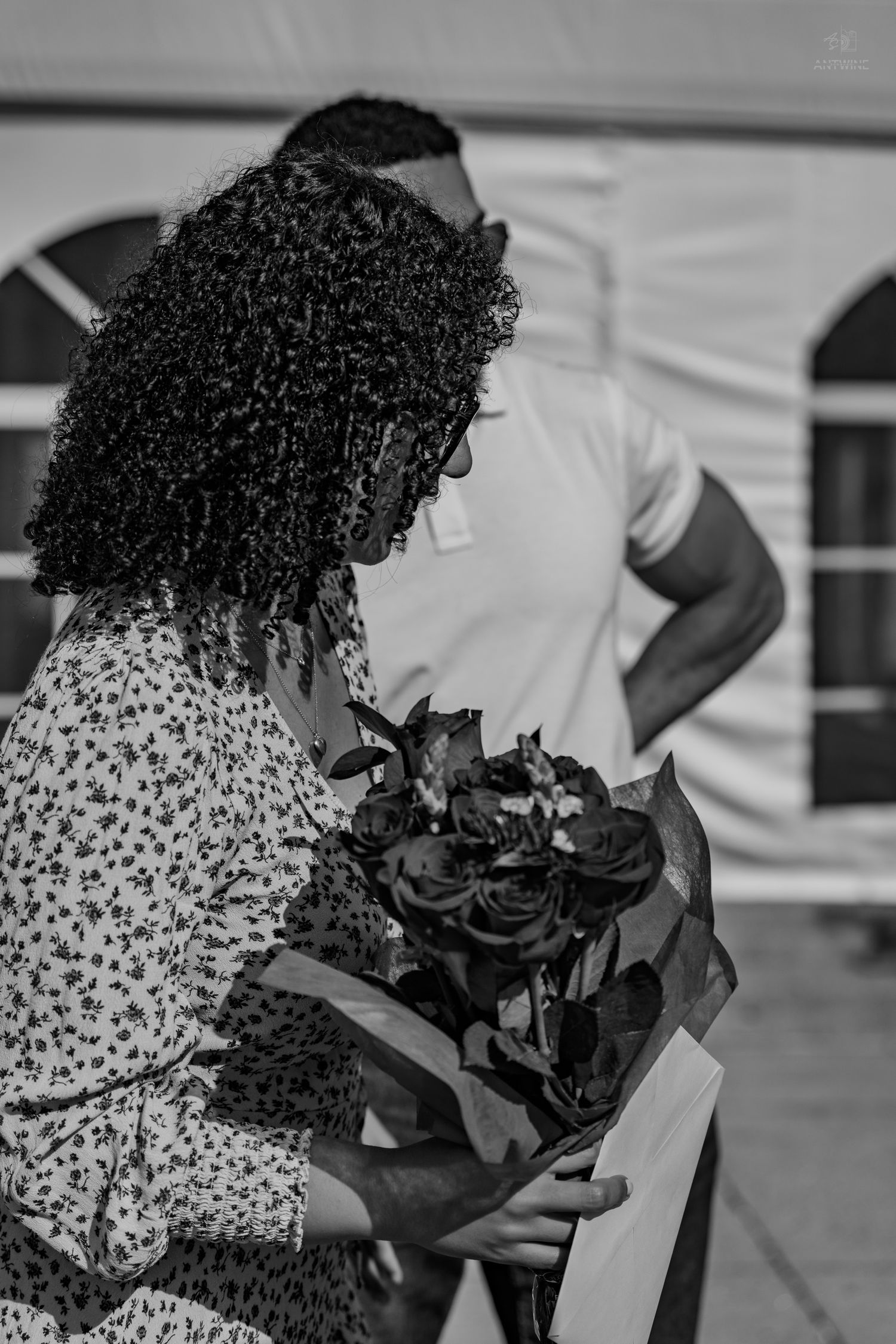 Black and white photo of a curly-haired woman holding roses while a man stands behind her near a tent.