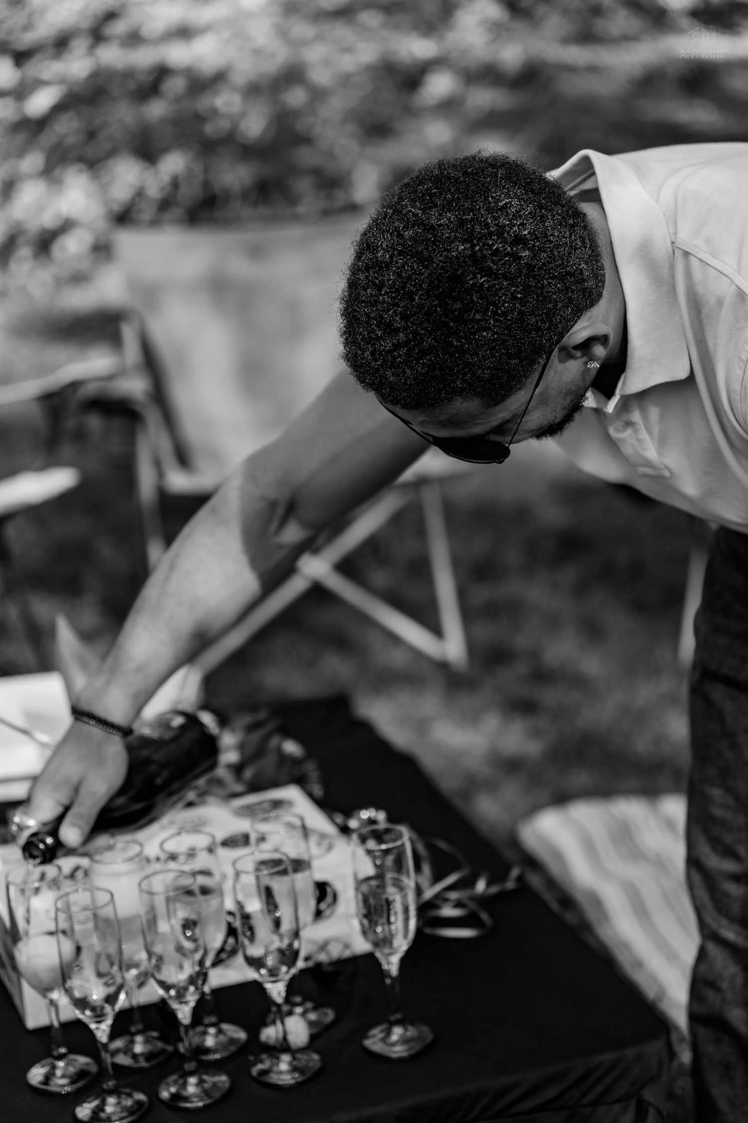 Man pouring champagne into flutes at an outdoor event table in black and white.