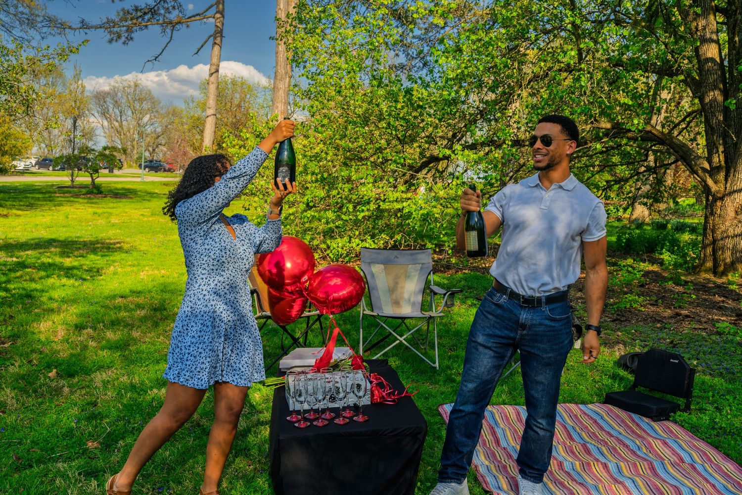 Happy couple toasting with champagne bottles near red heart balloons during a romantic outdoor park picnic.