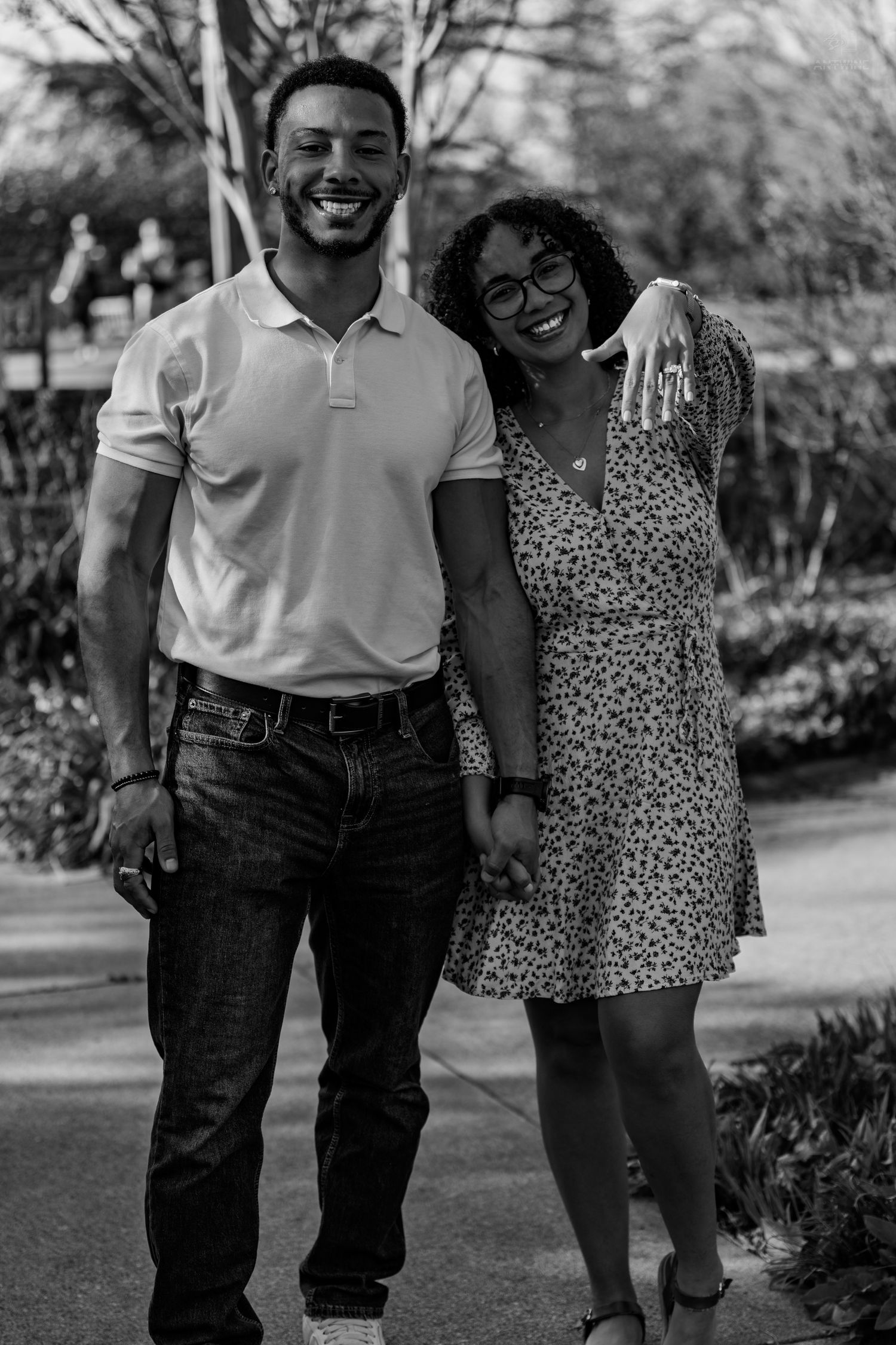 Black and white photo of a laughing couple embracing on a park path, the woman displaying an engagement ring.
