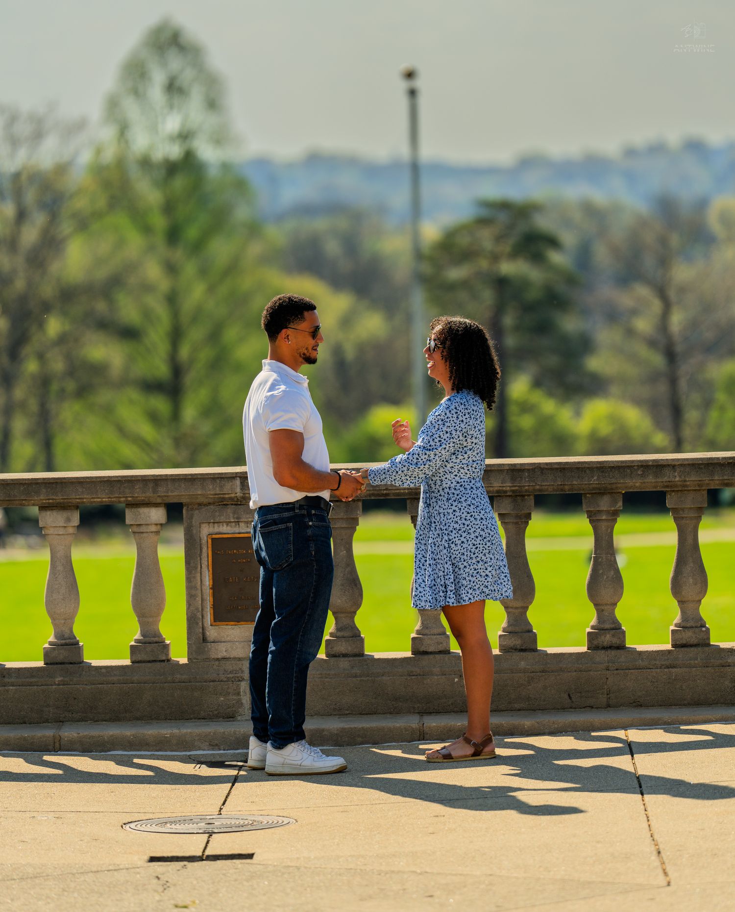 Couple smiling and holding hands on a bright sunny stone terrace with green park backdrop.