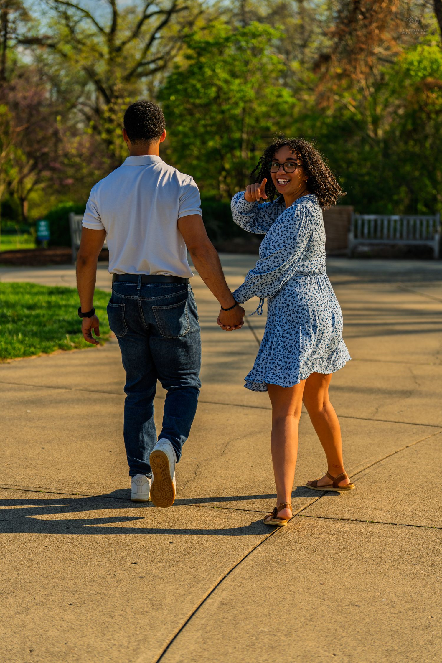 Woman in blue floral dress smiles back at camera while walking hand in hand with partner in park.