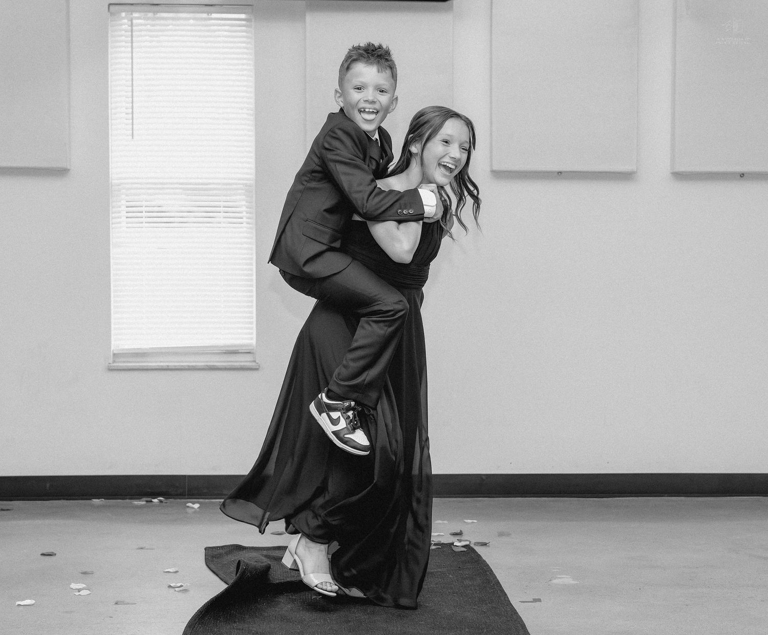 Black and white photo of two children in formal wear, one giving the other a piggyback ride indoors.