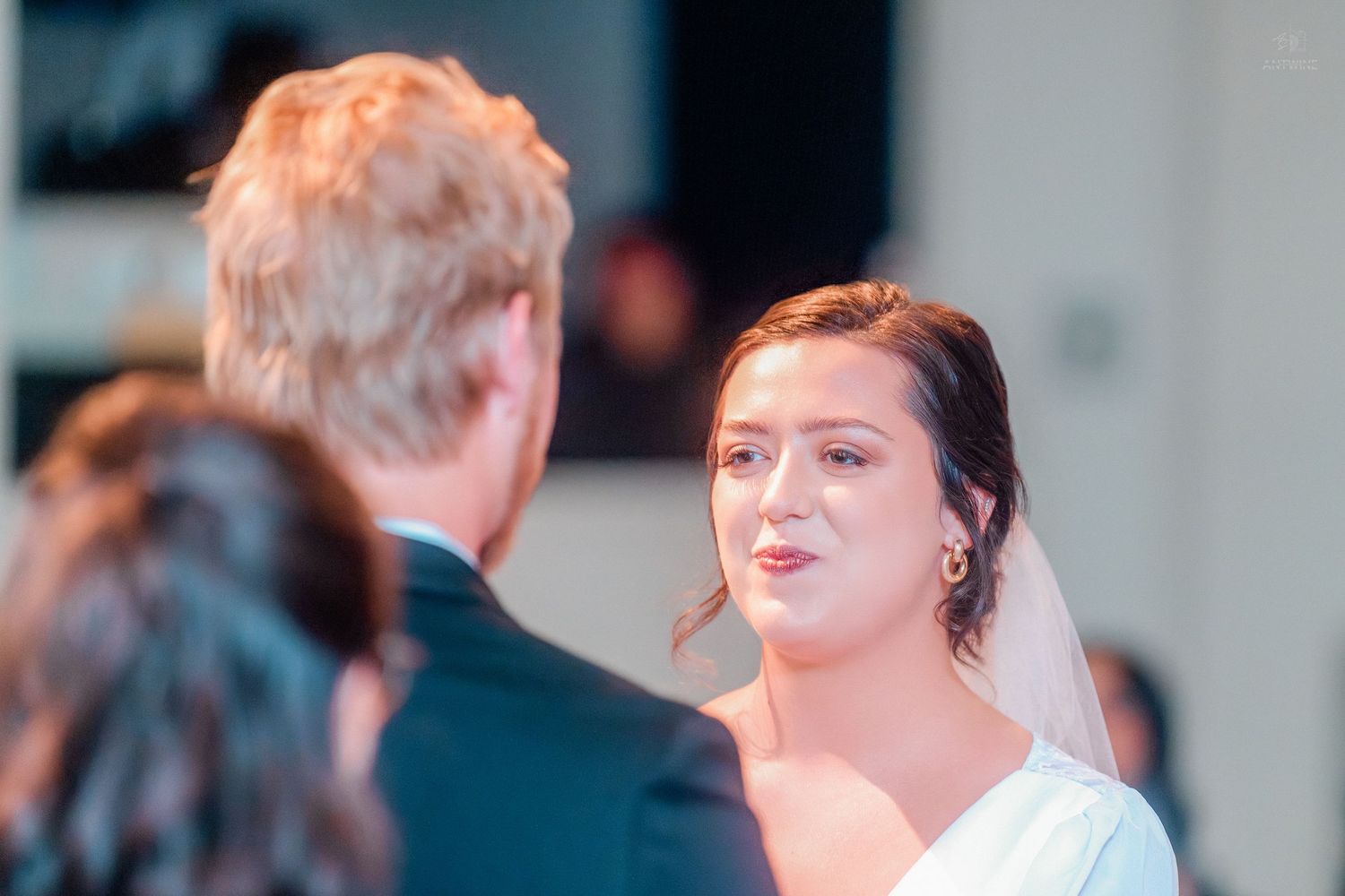 Bride in white veil gazes at officiant during emotional indoor wedding ceremony.