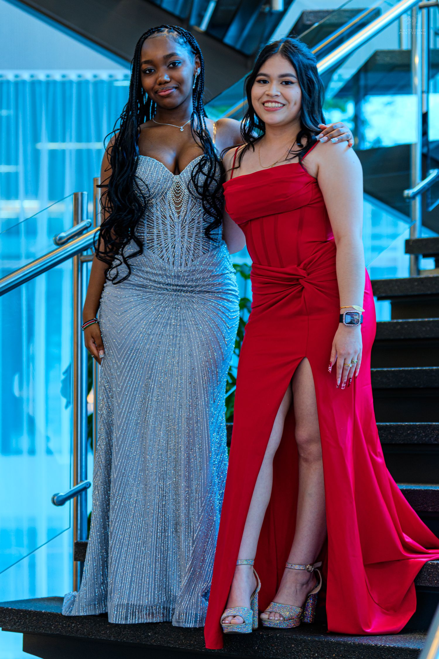 Two young women in elegant prom gowns, one silver beaded and one red slit dress, posing on stairs.