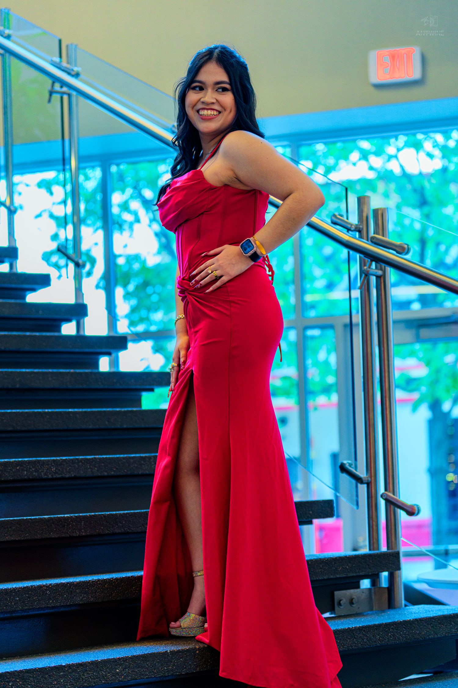 Woman in red one-shoulder slit gown posing on dark staircase in modern building.