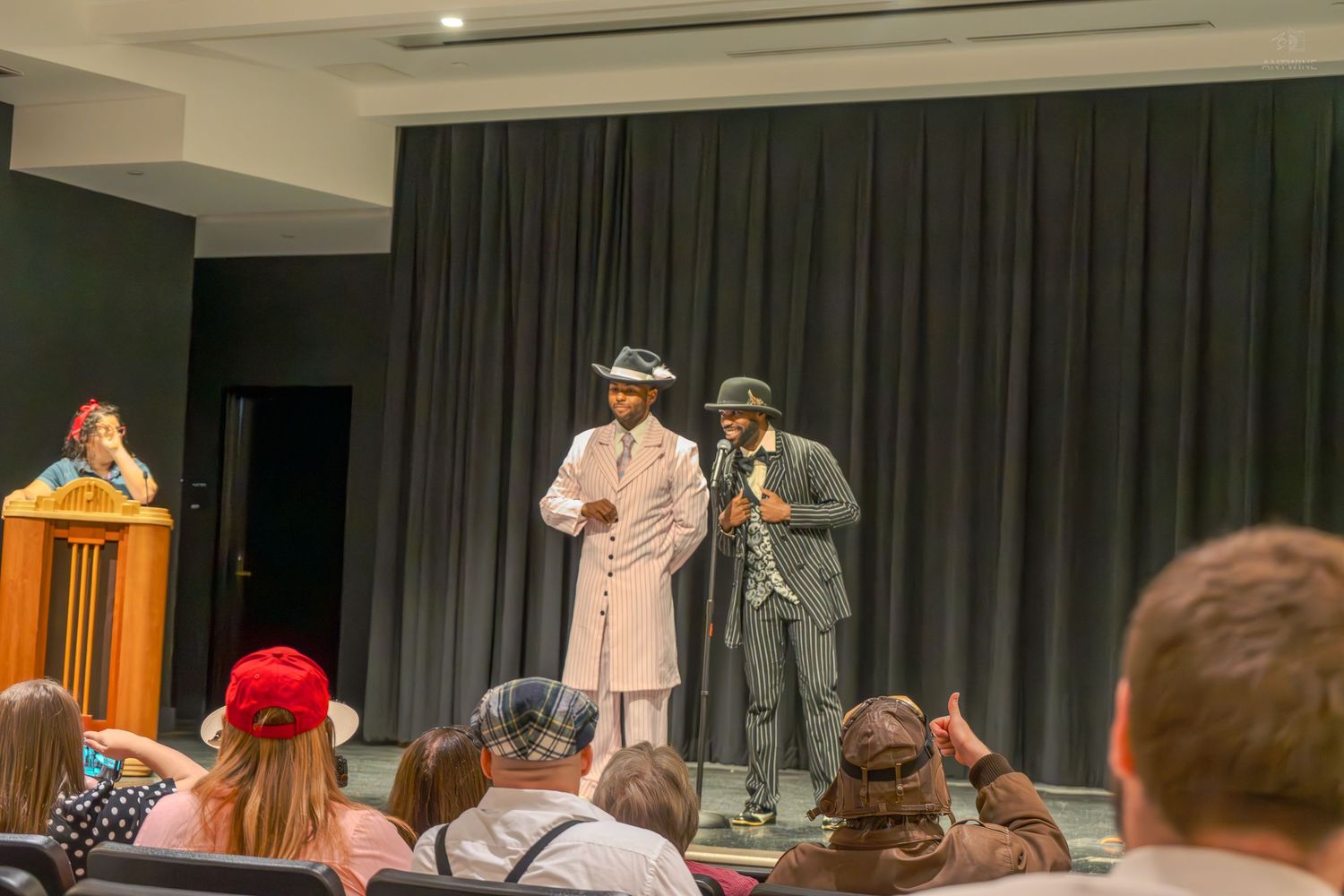 Two performers in fedora hats and suits on stage during a live theater show with an audience watching.