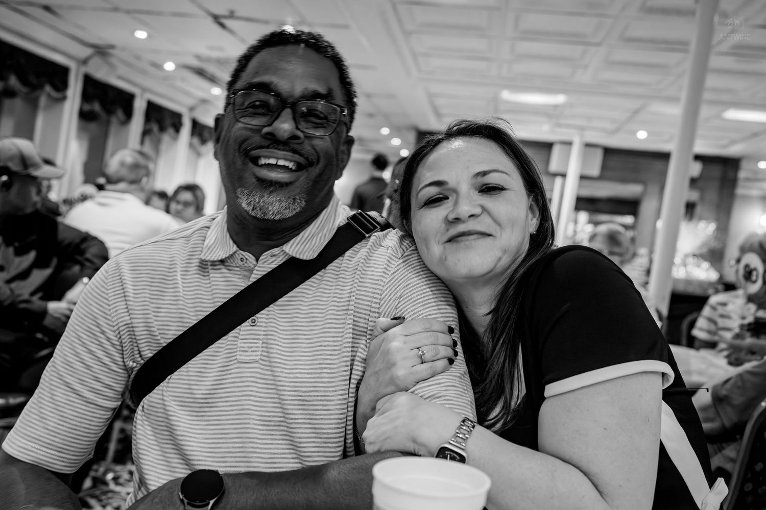 A smiling man and woman pose together at an indoor event, the woman holding a cup.