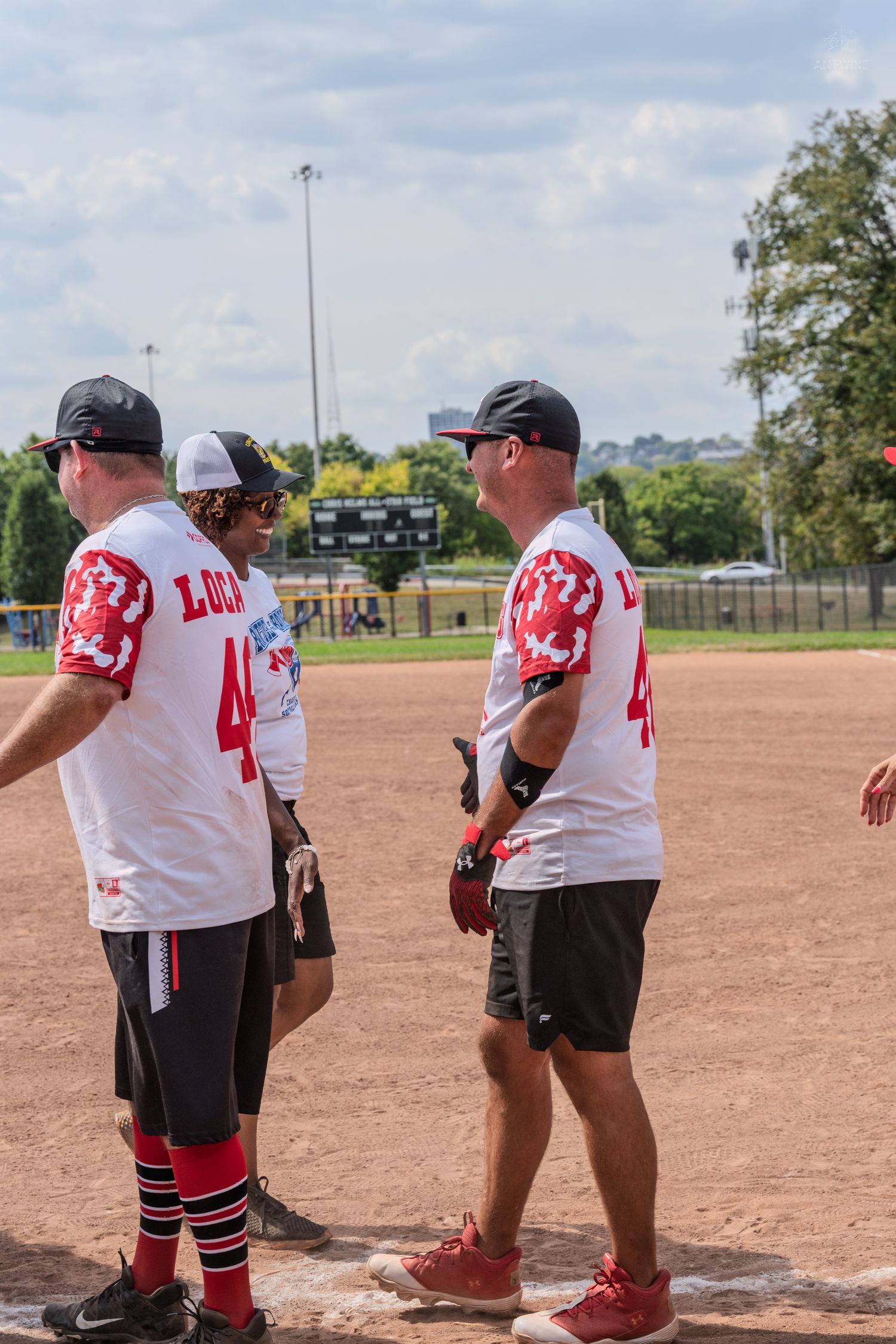 Three softball players in white LUCA jerseys and caps standing on base during an outdoor summer baseball game.