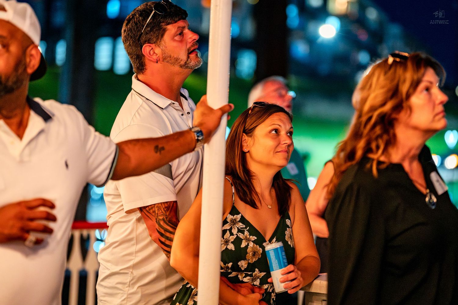 Group of adults watching an outdoor nighttime event, man in white polo shirt leaning on pole with tattoos visible.