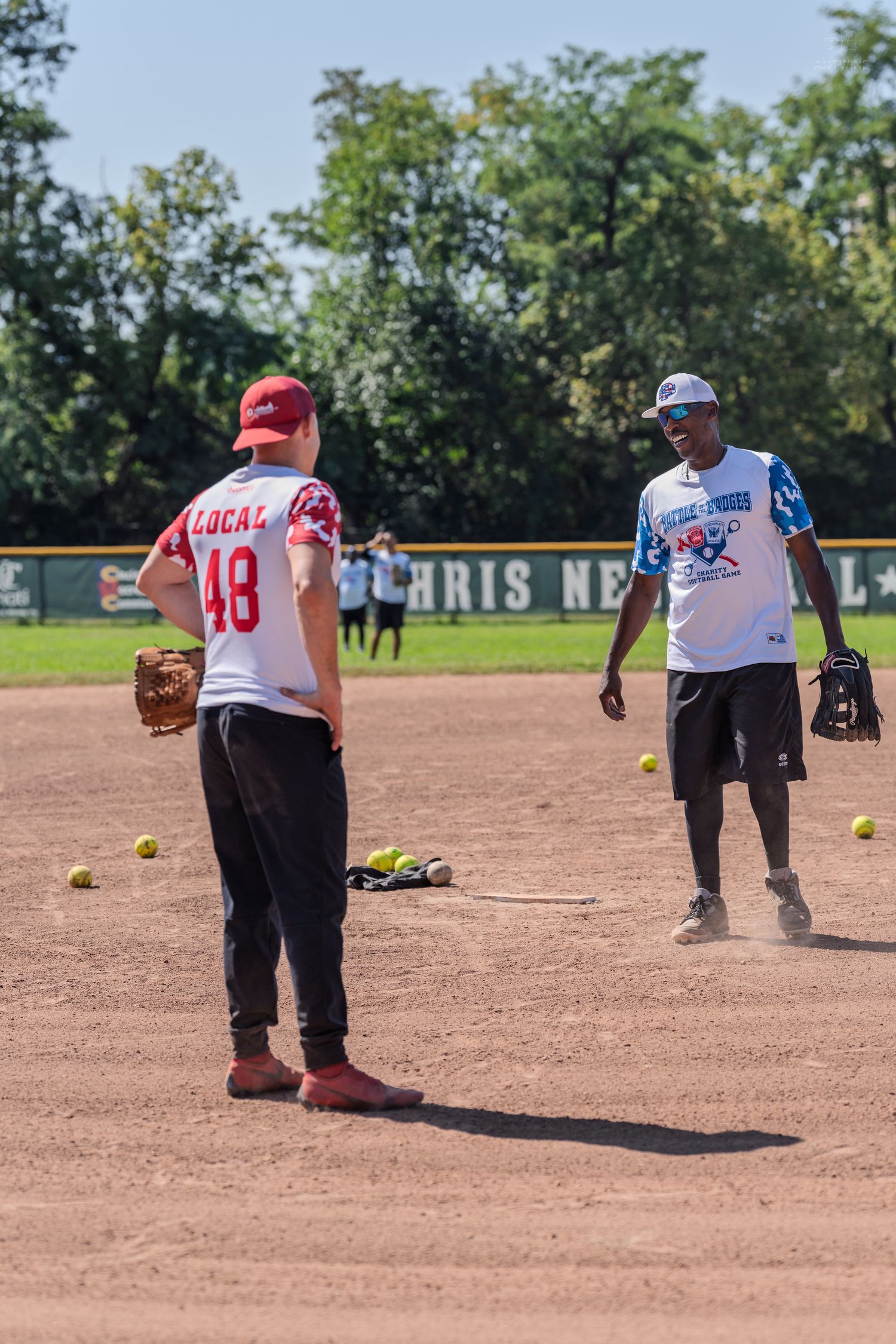 Two softball players practice fielding drills on a dirt infield during an outdoor game on a sunny day.