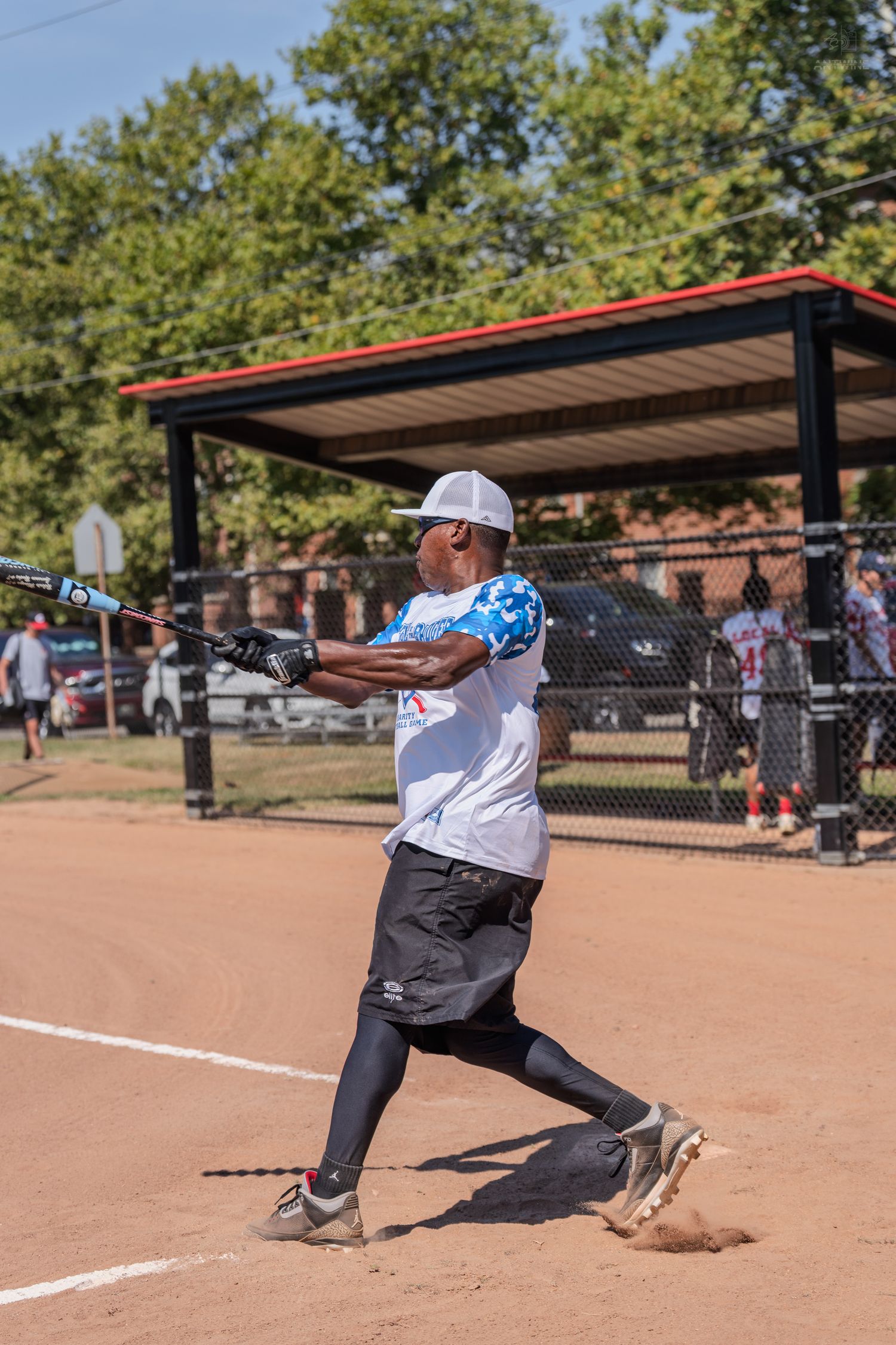 Male softball player in white jersey and cap completing a powerful swing at outdoor baseball field.