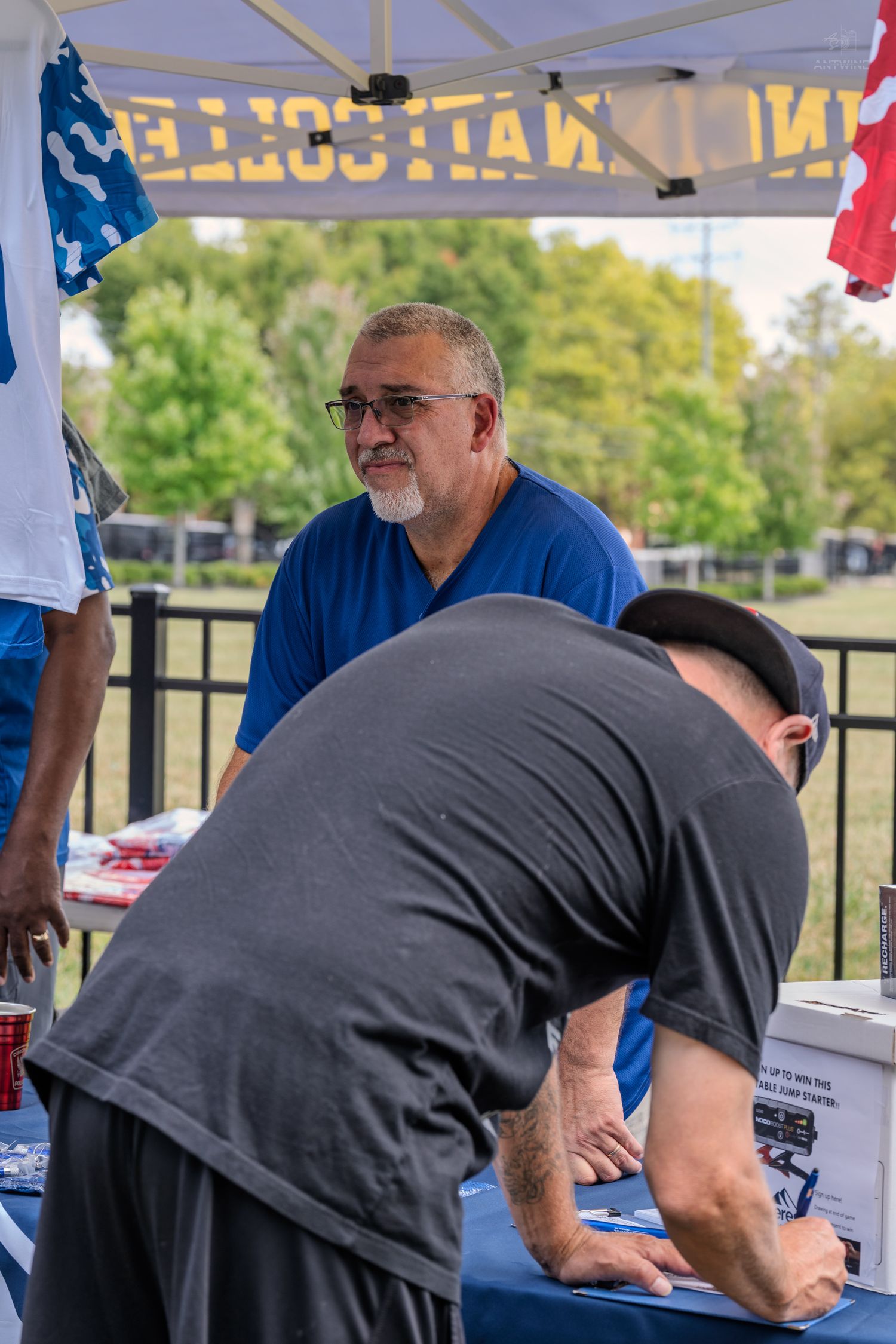 Man in blue shirt at registration table under tent at outdoor charity softball event.