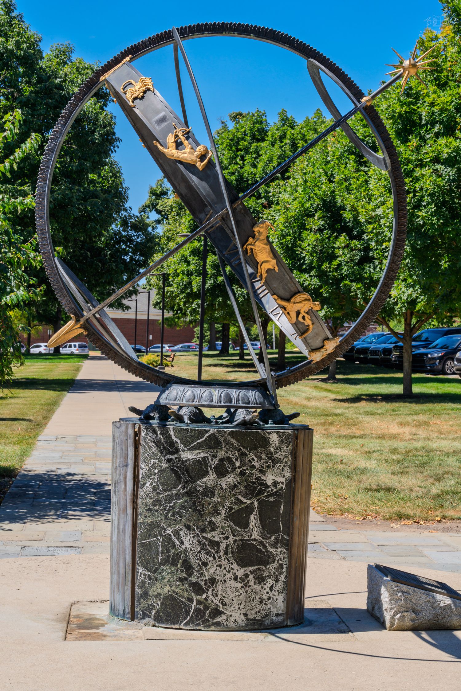 Ornate metal armillary sphere sundial on marble pedestal in a sunny park with green trees and pathway.