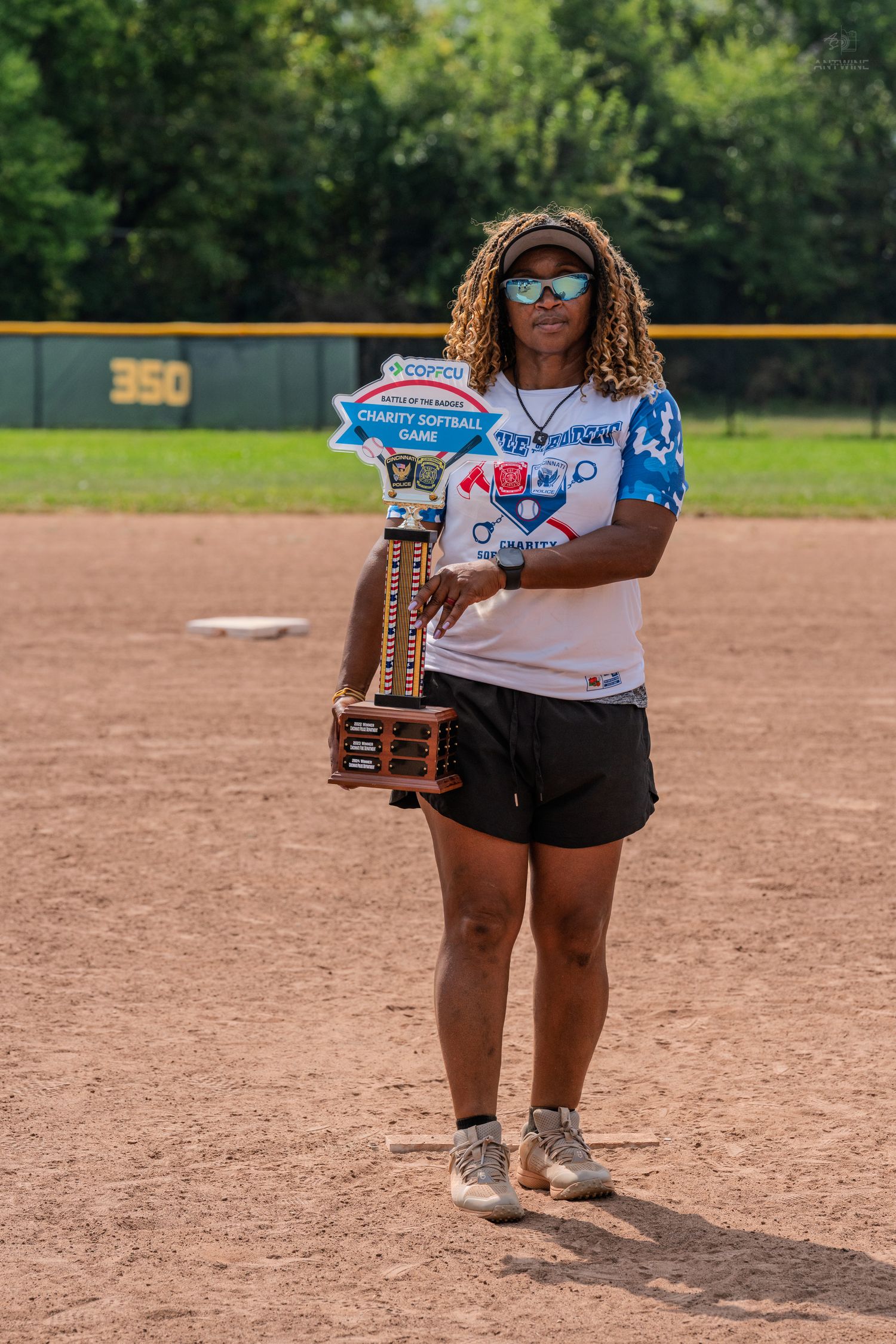 Woman in Battle of the Badges softball shirt holds a large championship trophy on a baseball field.