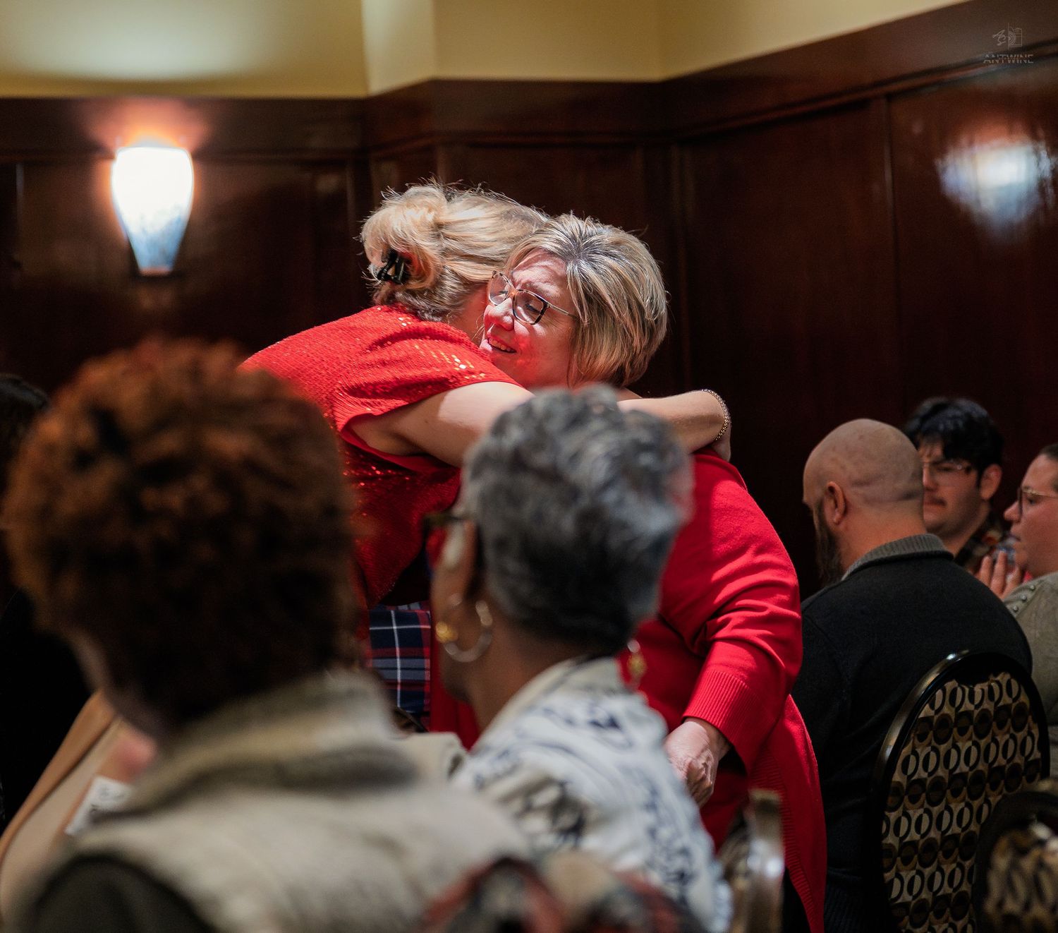 Two women in red embrace warmly at a social gathering in a wood-paneled room.