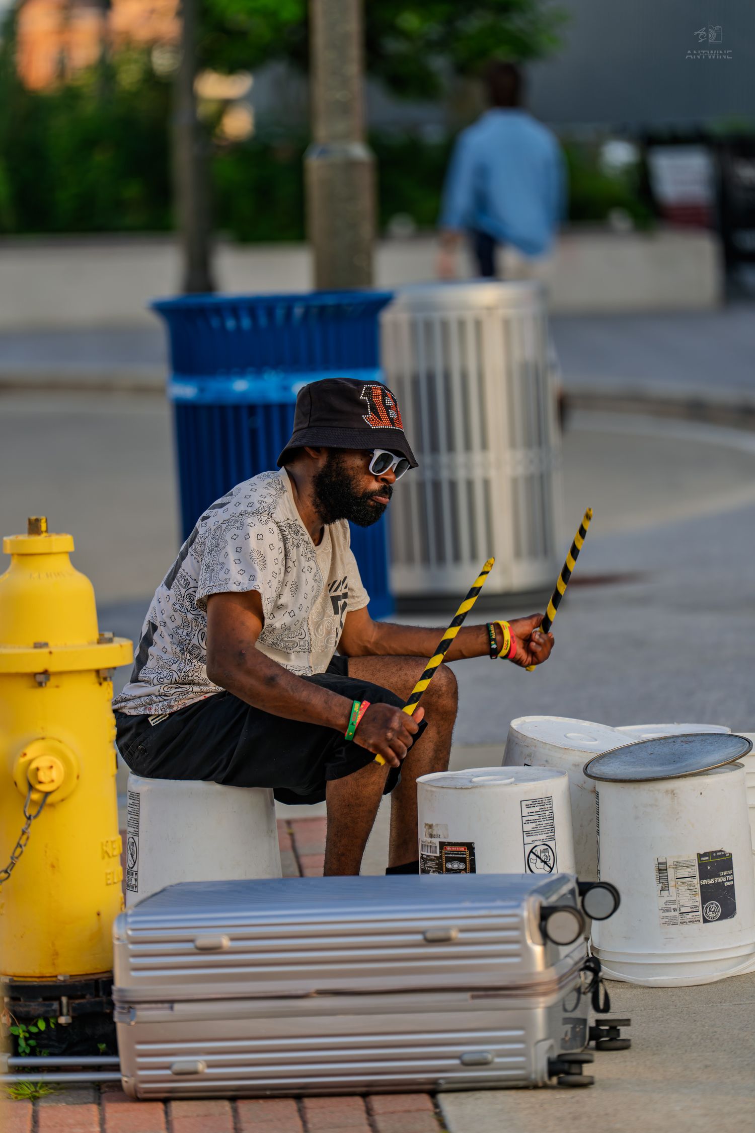 Street musician playing bucket drums with yellow-striped sticks near a fire hydrant in an urban setting.