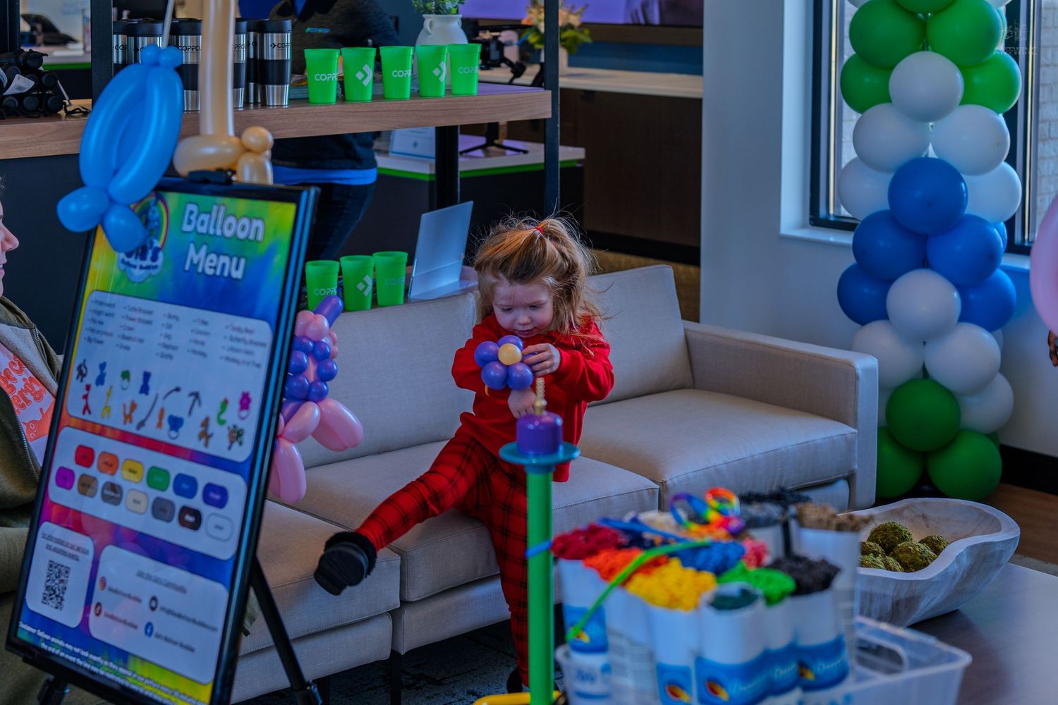 A toddler in red sits on a couch surrounded by colorful balloons and a balloon menu sign at a festive event.