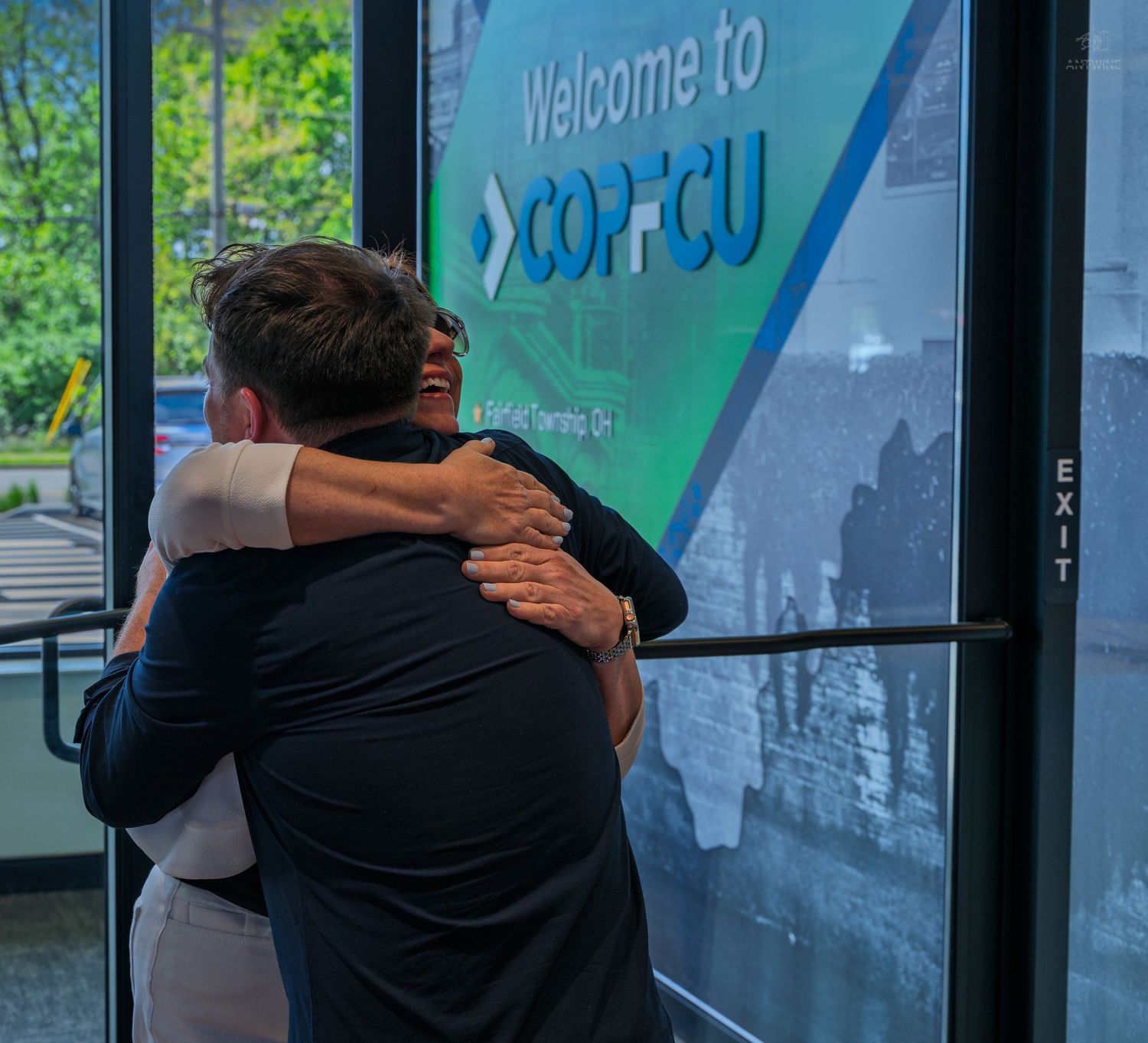 Two people share a warm hug in front of a Welcome to COPFCU credit union sign near an exit door.
