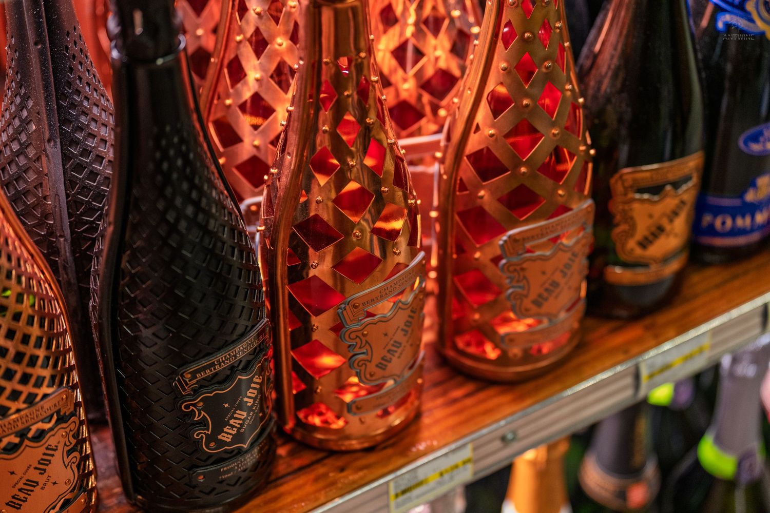 Ornate wine bottles with decorative gold lattice cages displayed on a shelf under warm red lighting.