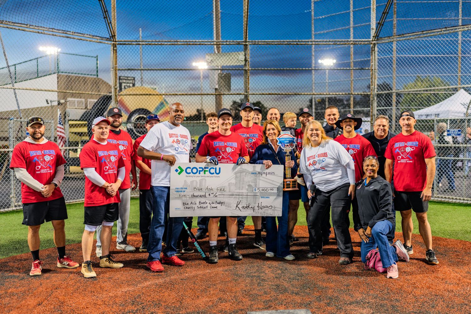 Softball team celebrates championship win holding large check and trophy under stadium lights at night.