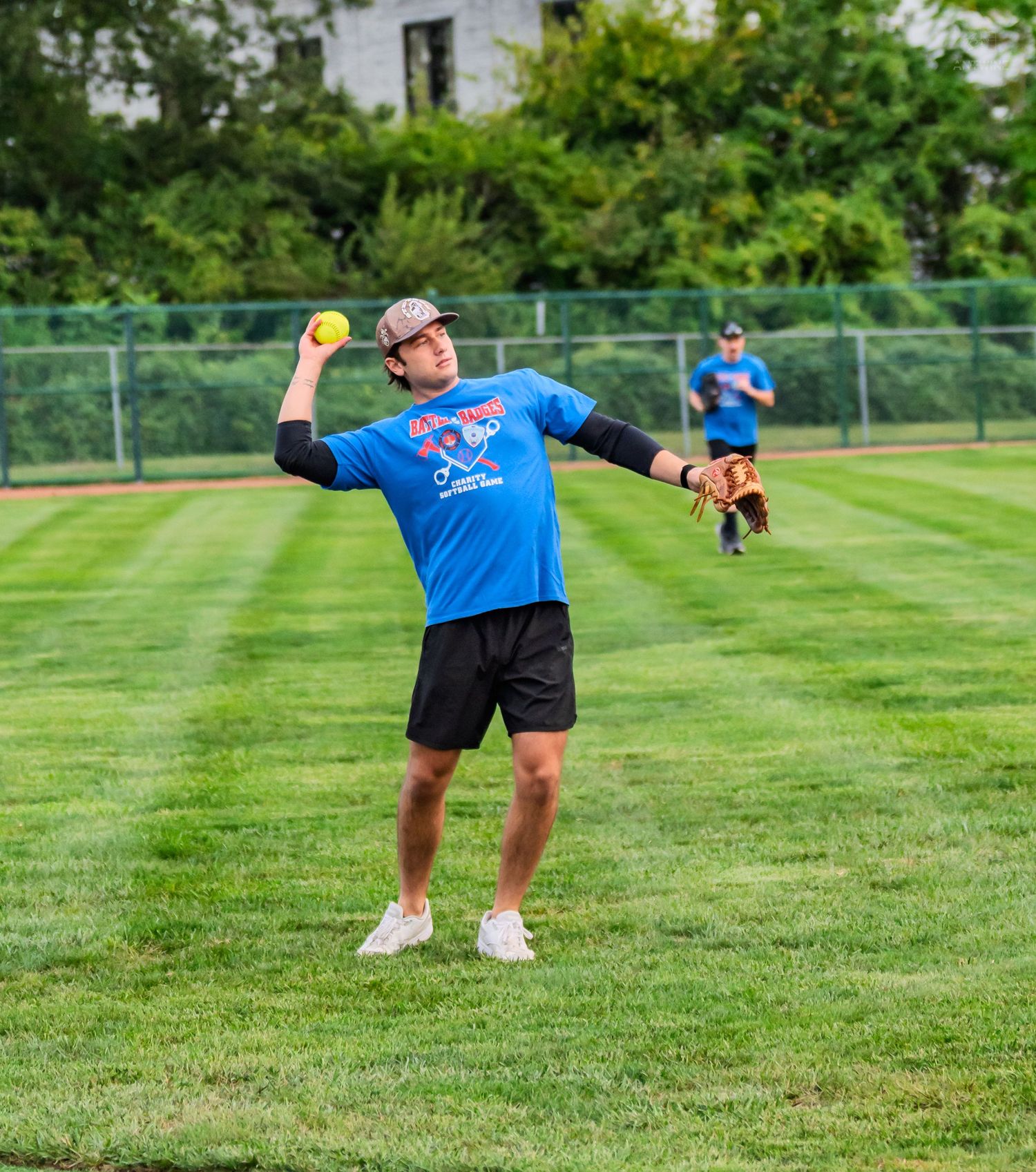A man in a blue shirt throws a yellow softball on a green grass field during an outdoor game.