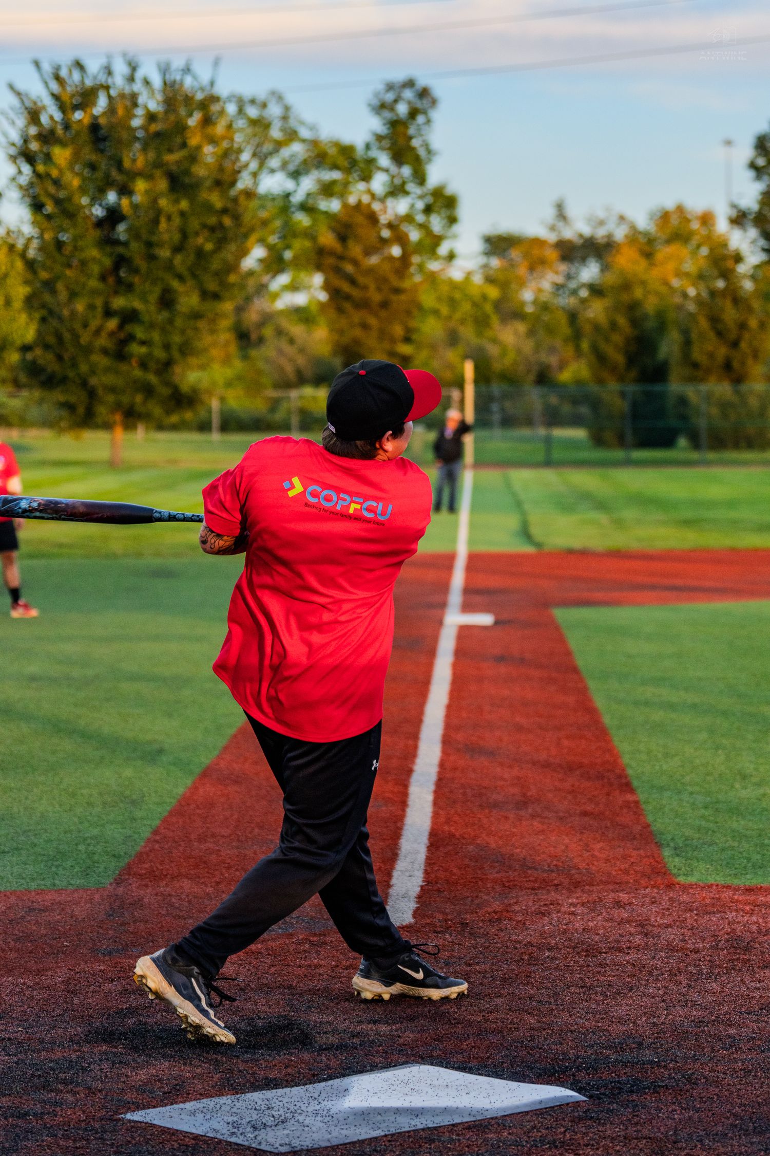 A batter in a red COPFCU shirt swings at a softball on a green field during a sunny evening game.