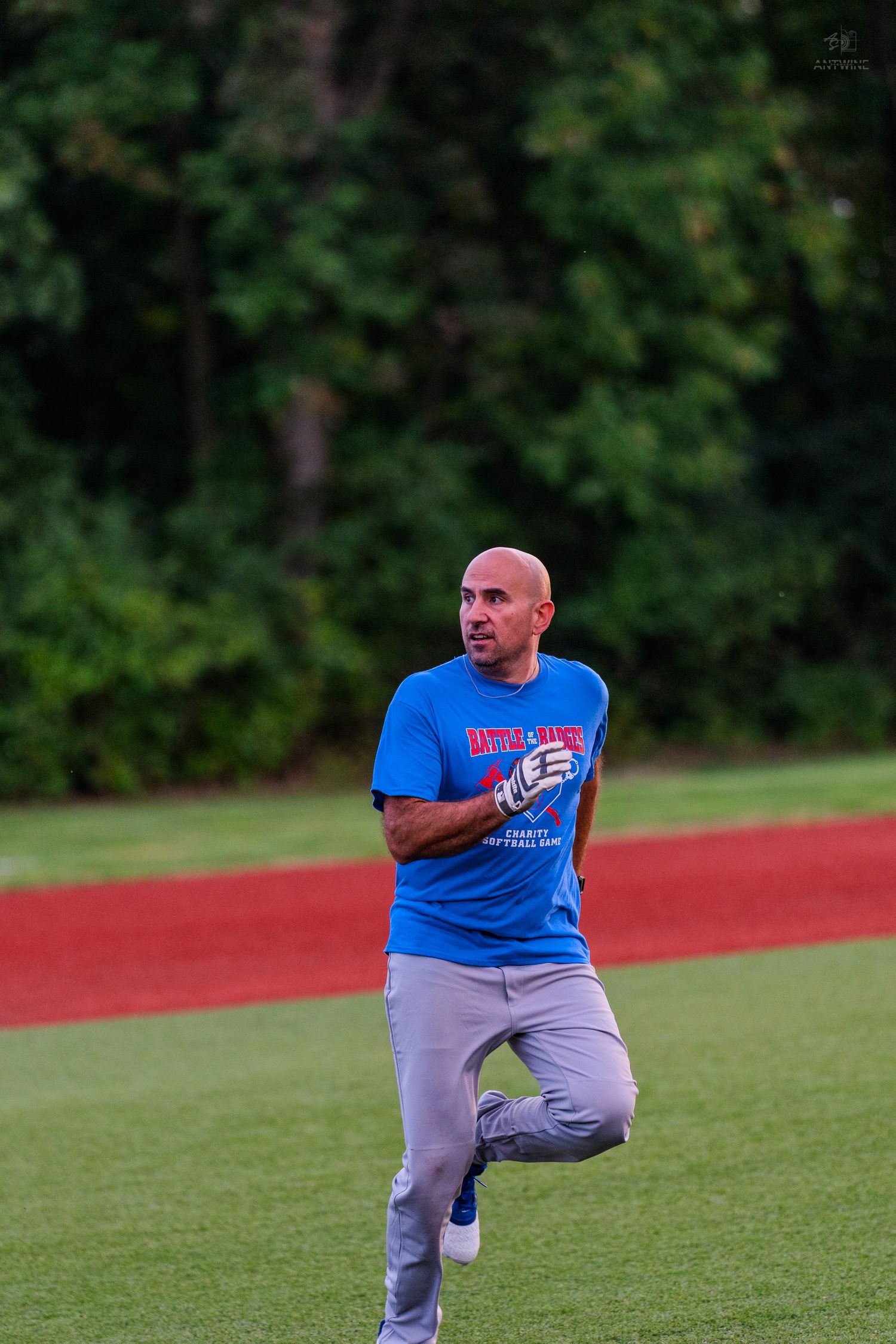Bald man in blue football t-shirt running on a green sports field with red track and trees in background.