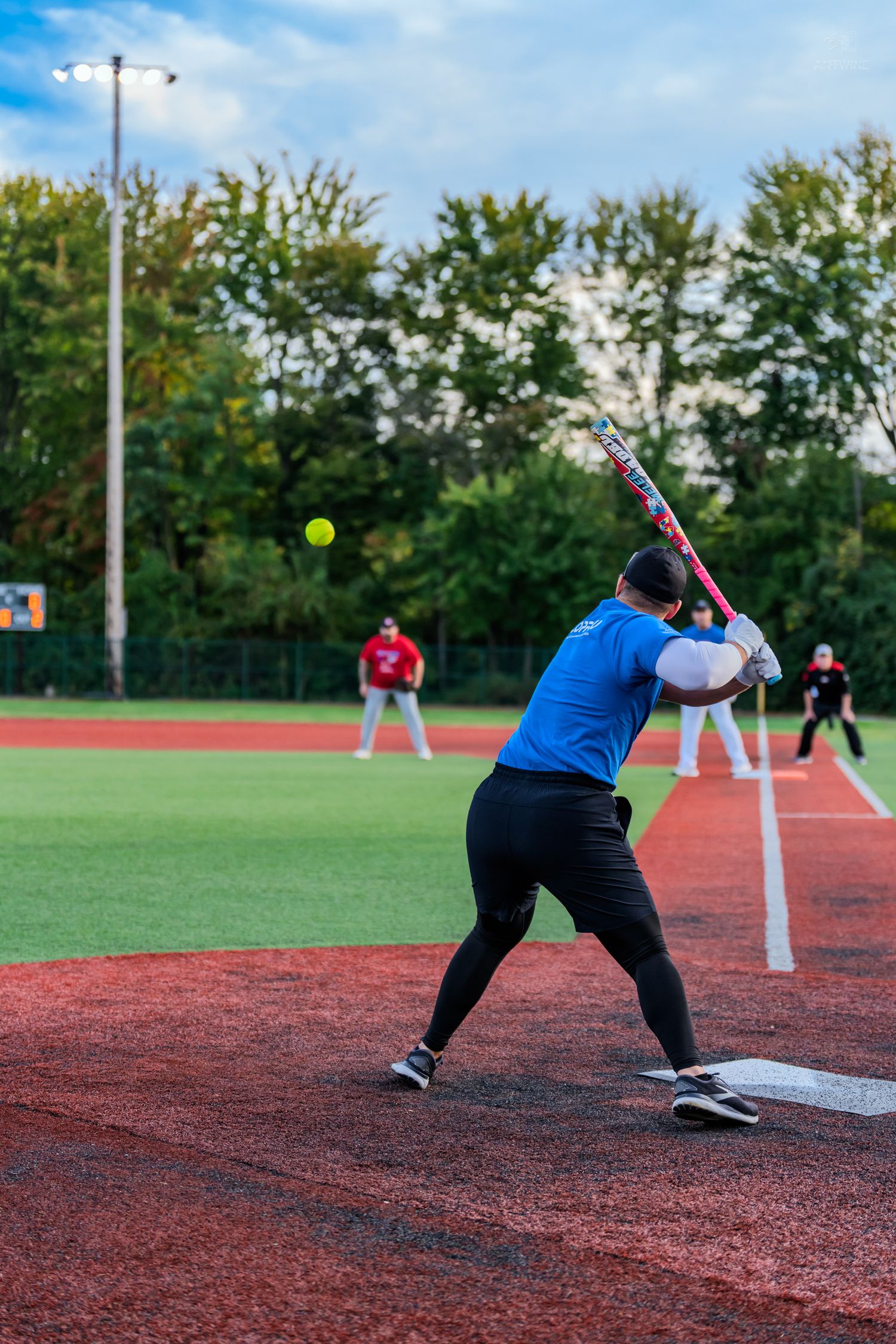 Batter in blue shirt swings at yellow softball on red turf field with outfielders in background.
