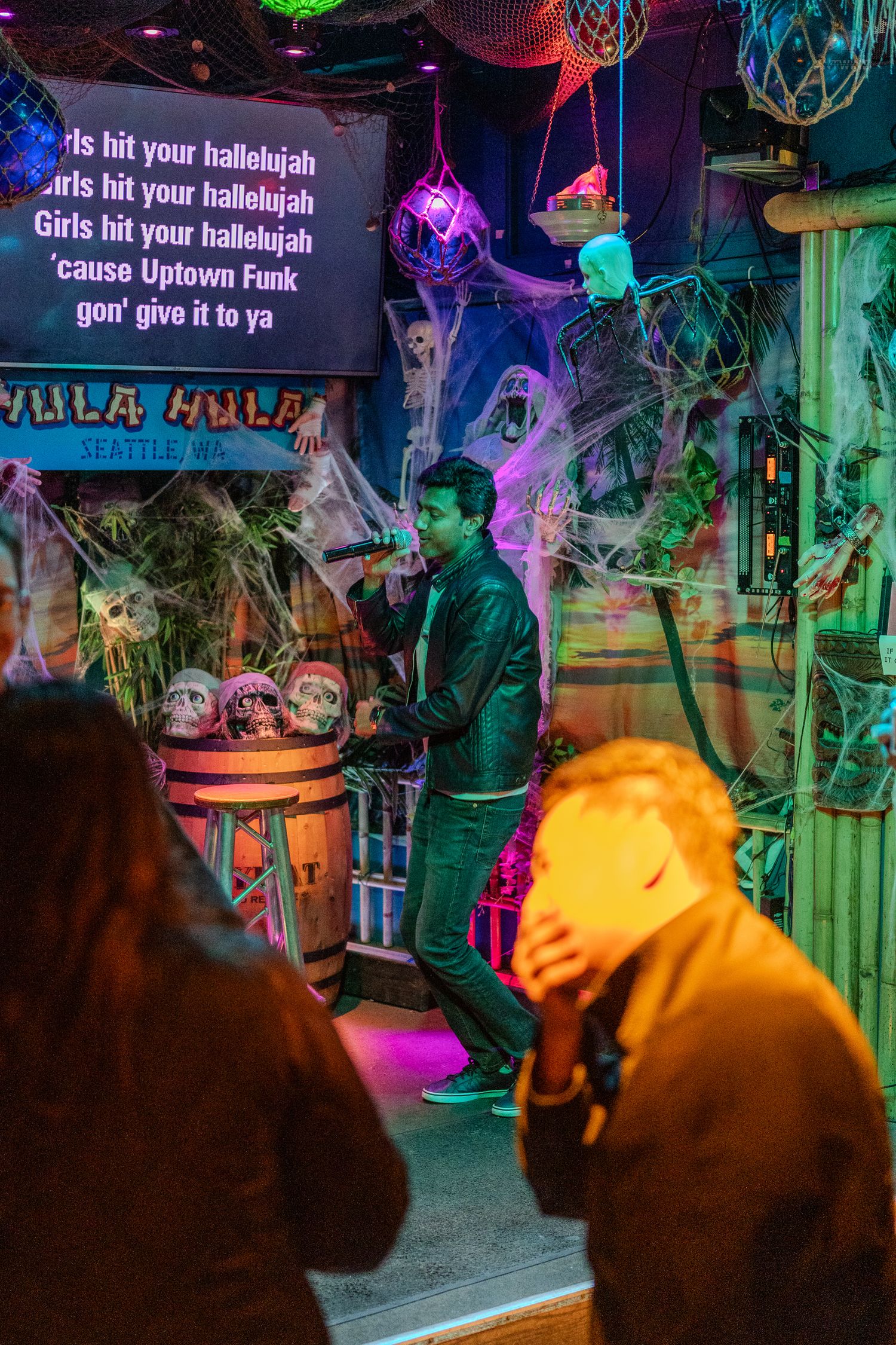 Man performing karaoke at Halloween-decorated Hula Hula bar singing Uptown Funk surrounded by skulls and spiderwebs.