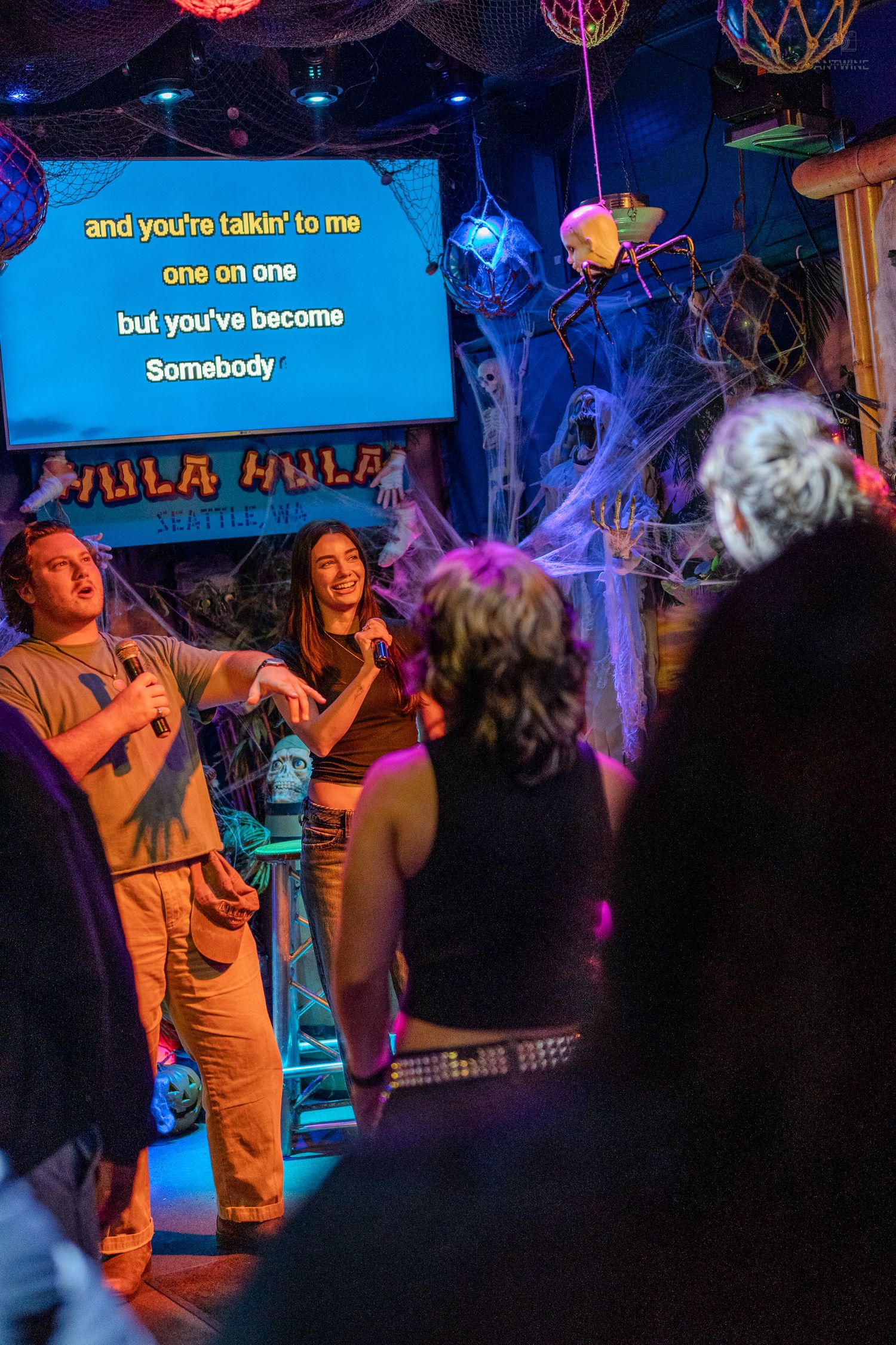 Duo performing Somebody karaoke at Halloween-themed Hula Hula Seattle bar with cobweb decorations.
