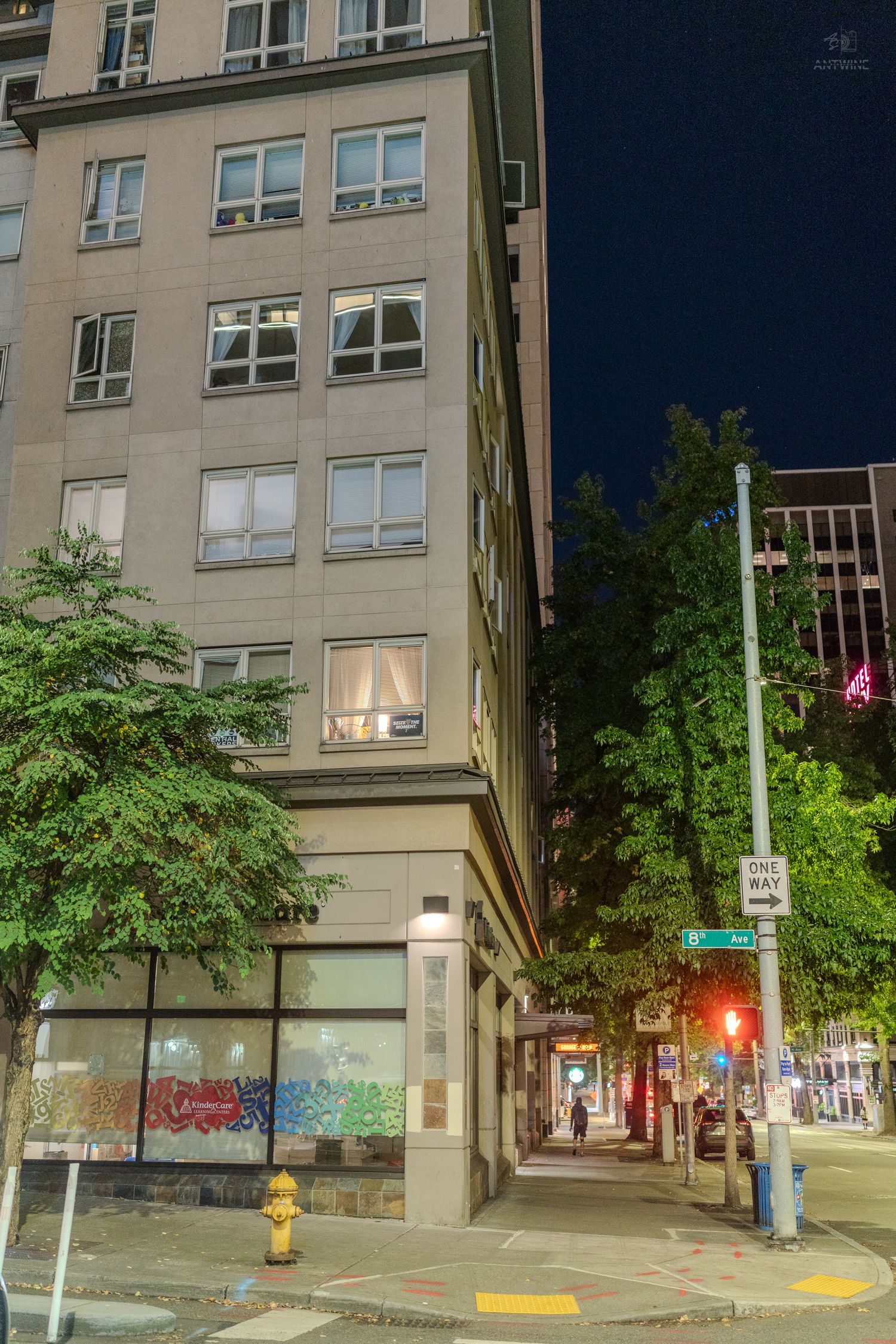 Nighttime urban street corner with a multi-story building, trees, fire hydrant, and illuminated storefronts in a city downtown.