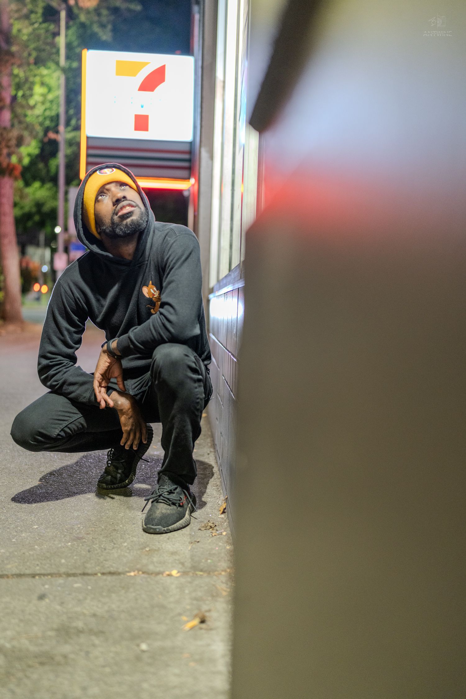 Man in black hoodie and yellow beanie crouching outside a 7-Eleven store at night.