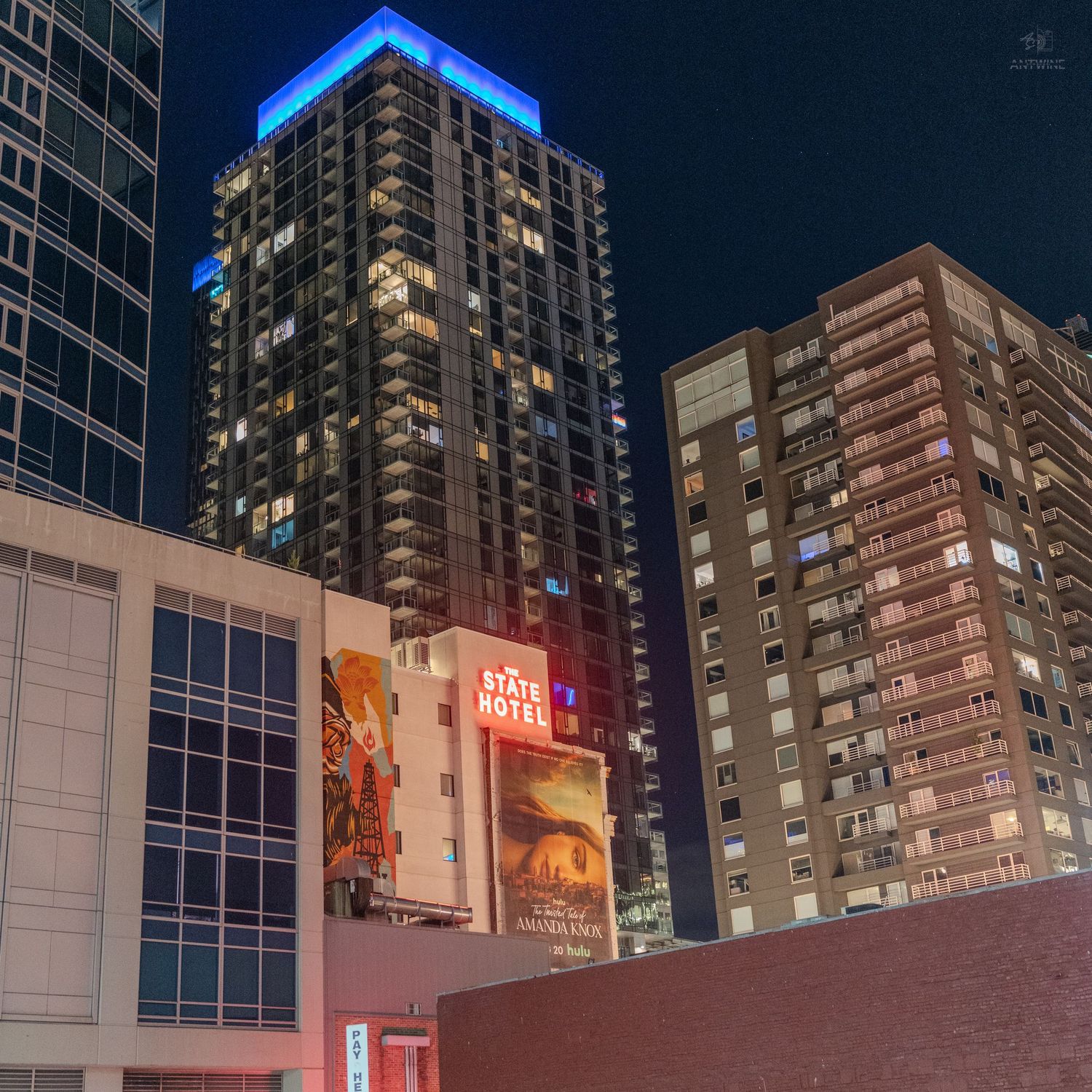 Nighttime cityscape with illuminated skyscrapers, a blue-lit rooftop, and a State Hotel billboard in an urban setting.