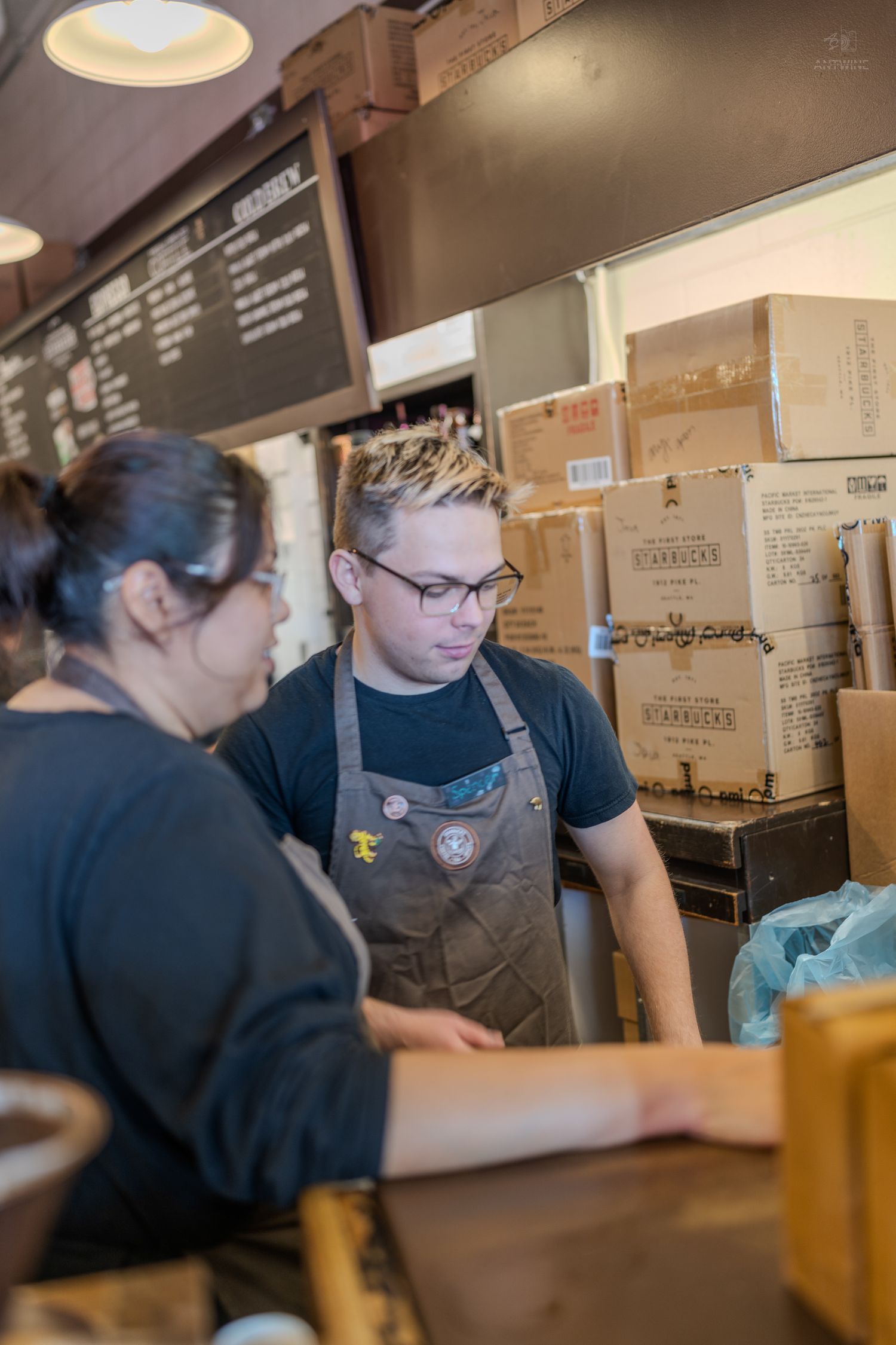 Two Starbucks baristas in brown aprons work together behind the counter with cardboard boxes stacked in the background.