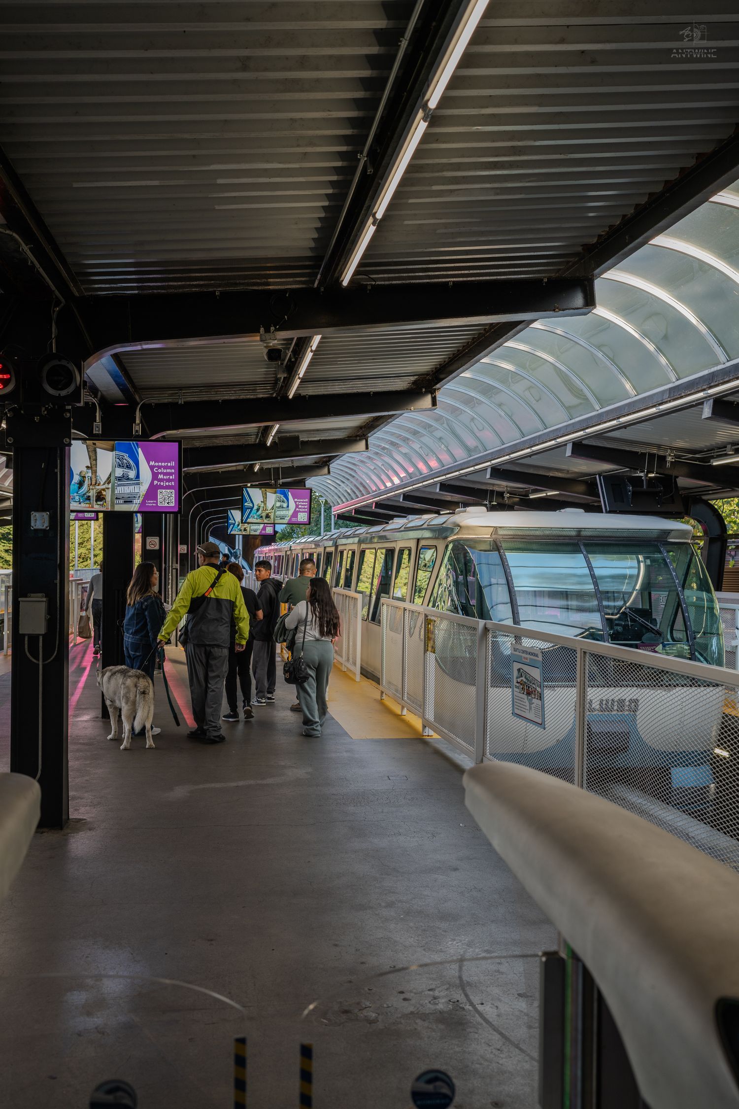 Passengers and a service dog wait on a covered Seattle monorail station platform beside a train.
