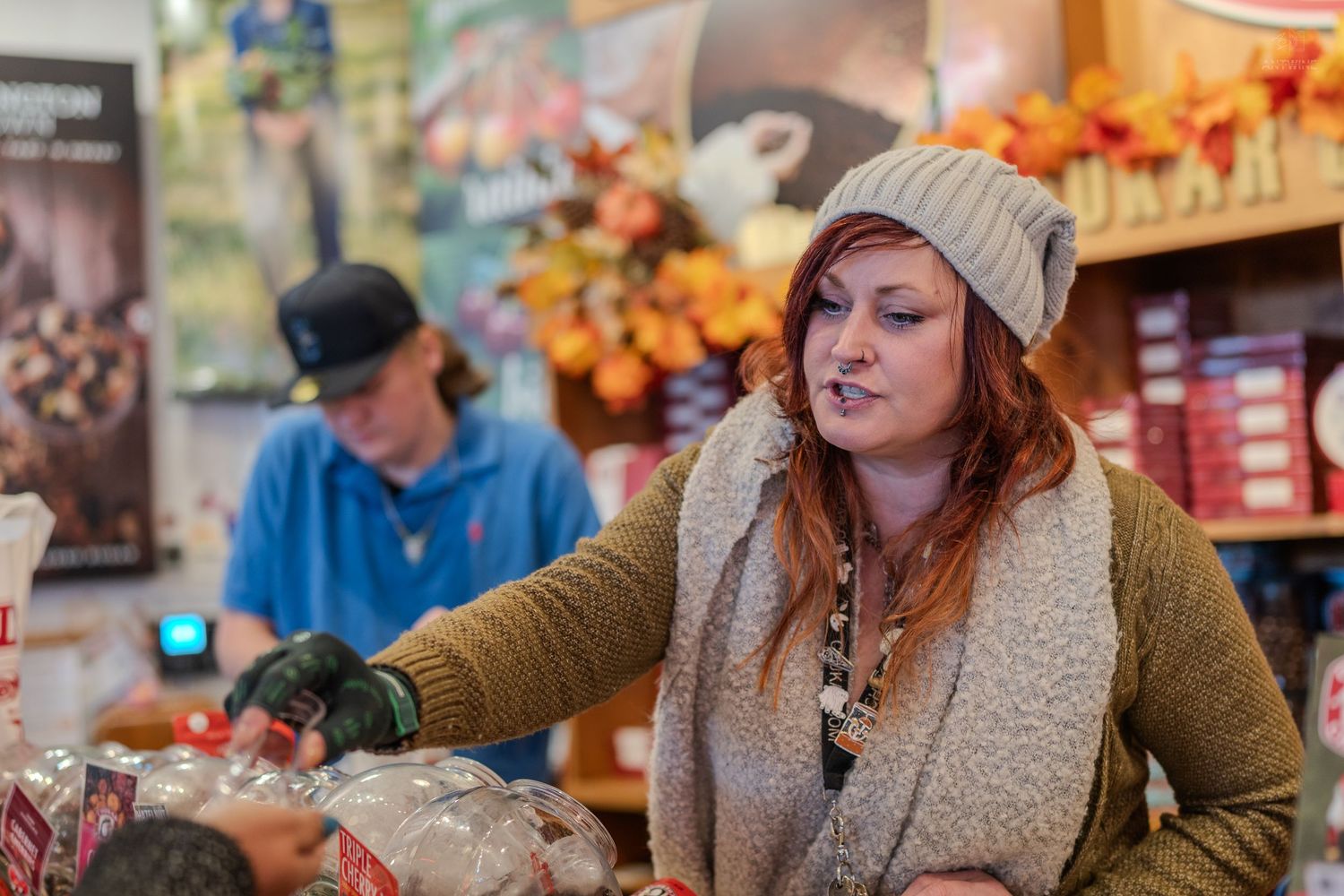 Woman in beige cardigan and gray beanie shopping at a fall-decorated store counter with another shopper in background.