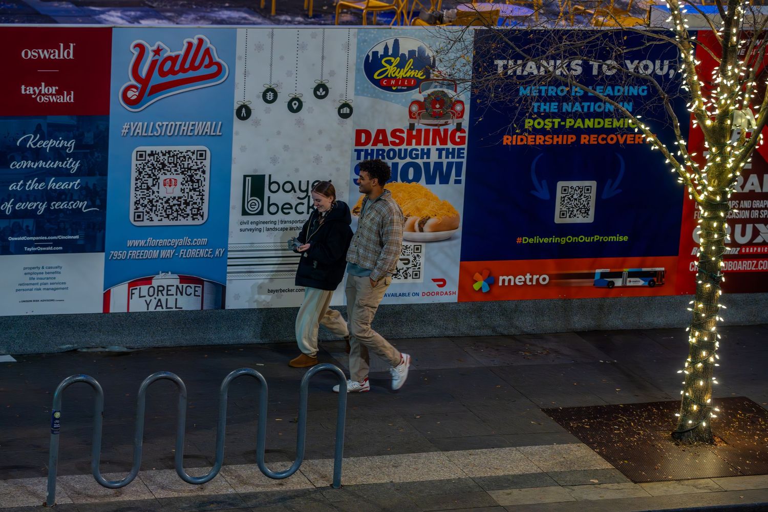 Two pedestrians walk past colorful urban billboards and holiday lights on a city street at dusk.